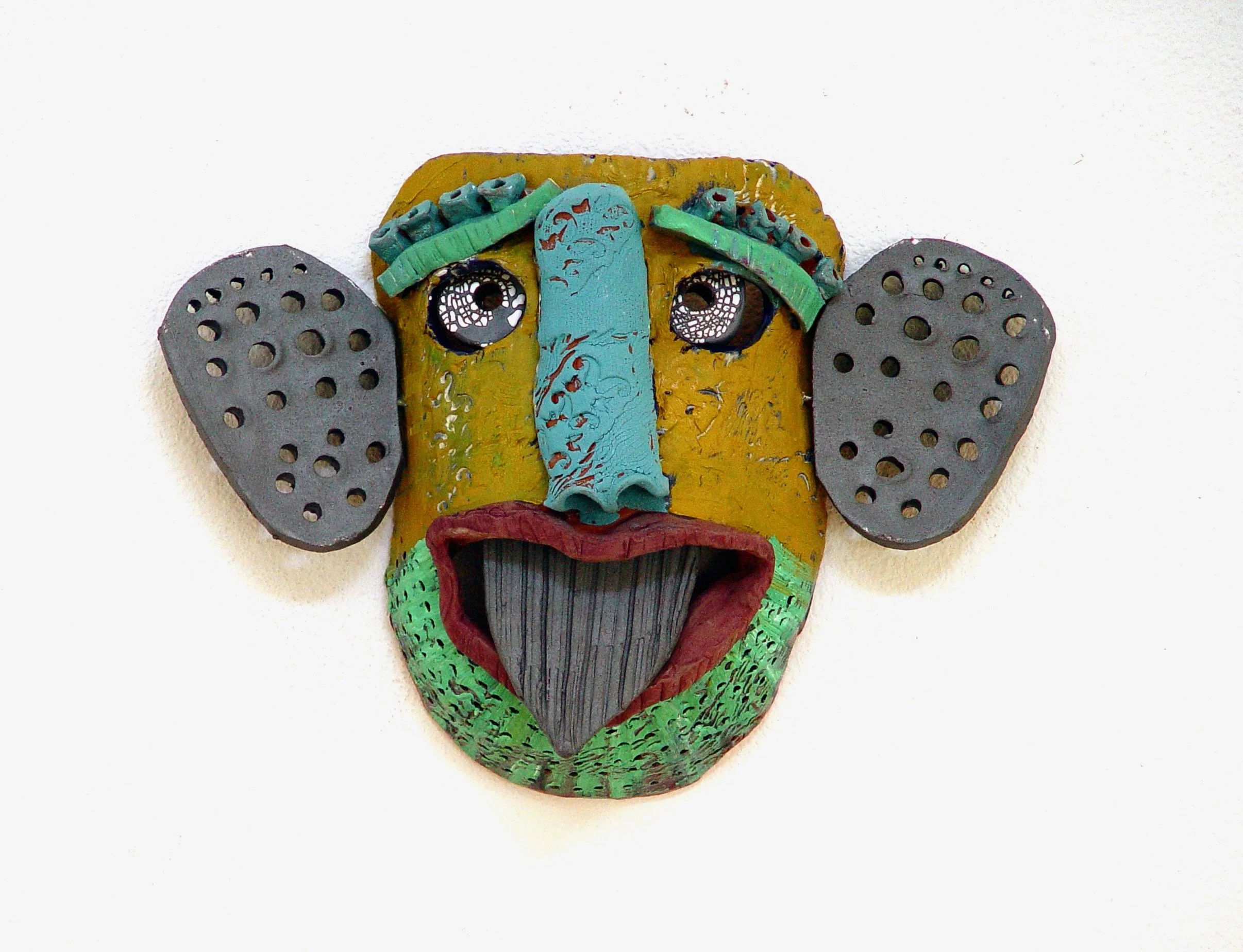 Colorful abstract face sculpture with large ears, prominent nose, and open mouth, mounted on white wall.