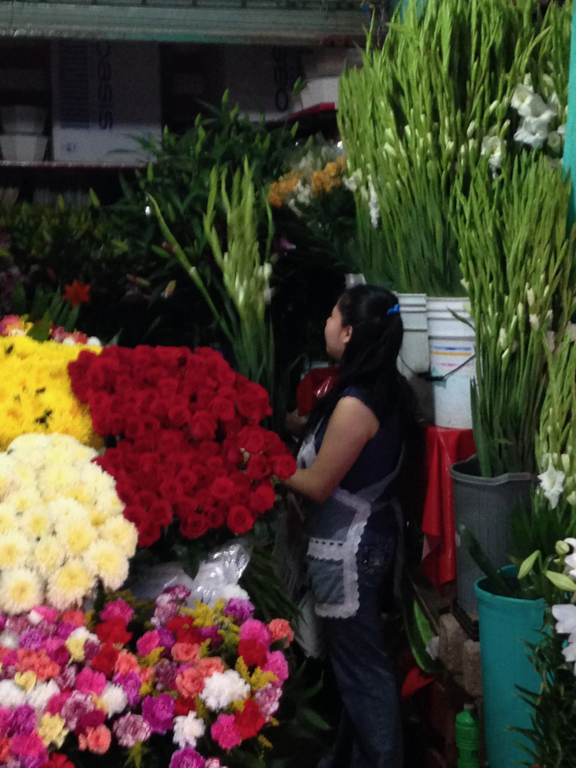 A woman shopping for flowers at a flower stand with various colorful flowers and green plants around her.