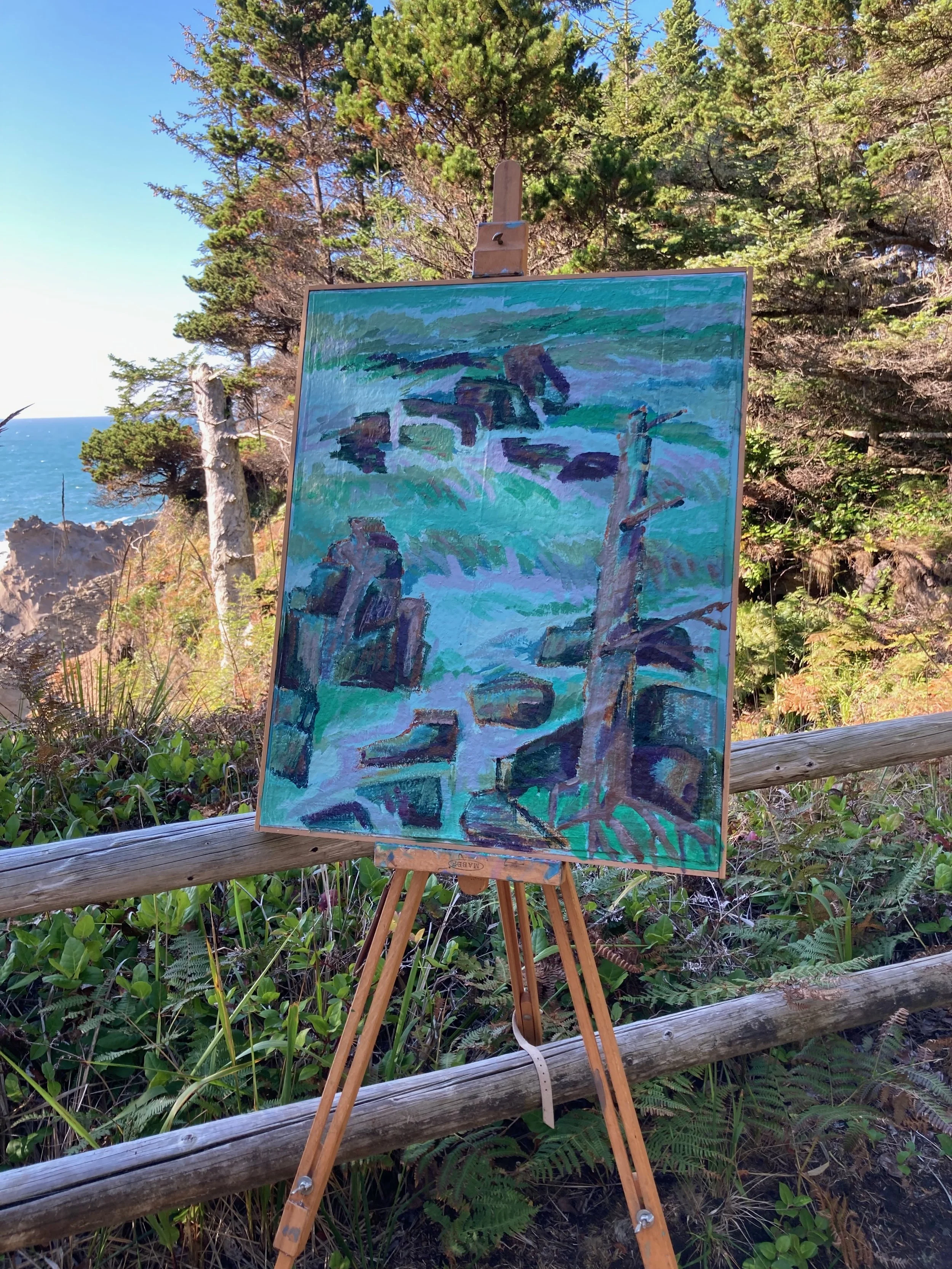 Landscape painting on an easel outdoors, featuring rocky formations and ocean waves, with trees and blue sky in the background.