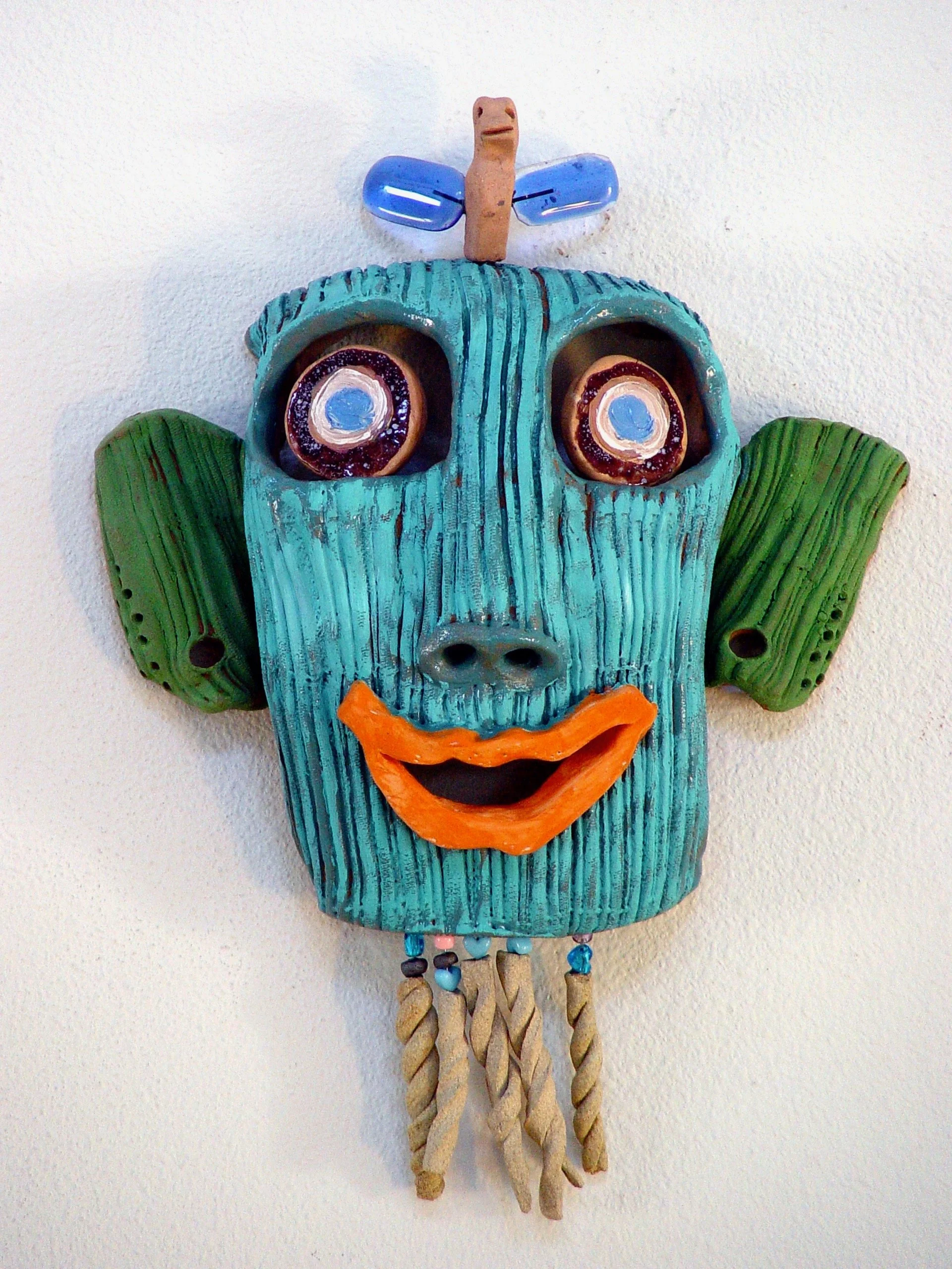 Colorful mask with exaggerated facial features including large eyes with spirals, green ears, a blue face with wood-like texture, a wide orange smile, and hanging rope and bead decorations, mounted on a white wall.