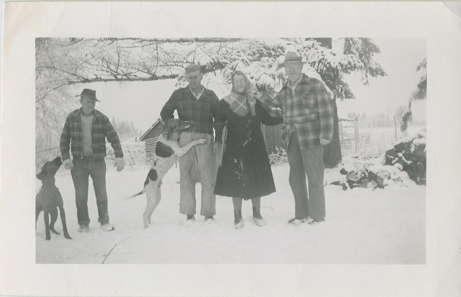 Black and white photo of four people and two dogs standing outside in the snow. The background shows snow-covered trees and a small building.
