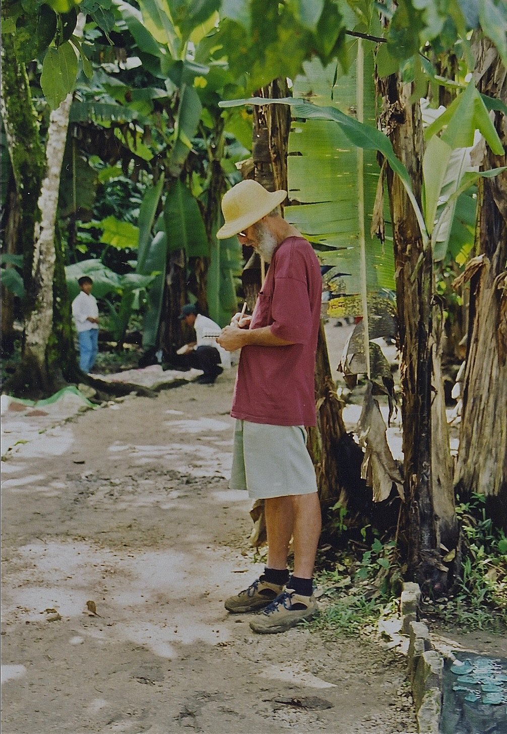 An elderly man with a beard, wearing a straw hat, red T-shirt, gray shorts, and hiking shoes, writing in a small notebook in a lush, green outdoor setting with banana trees. In the background, there are a few children and adults, some sitting and some standing, among the trees.