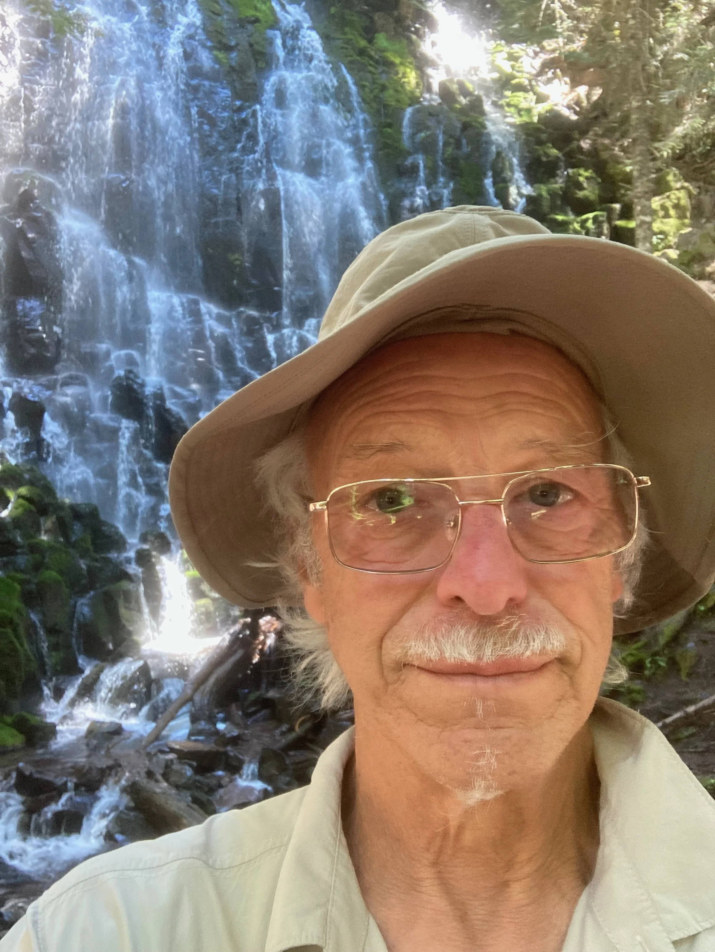 An elderly man with glasses and a beige hat taking a selfie in front of a waterfall in a forest.