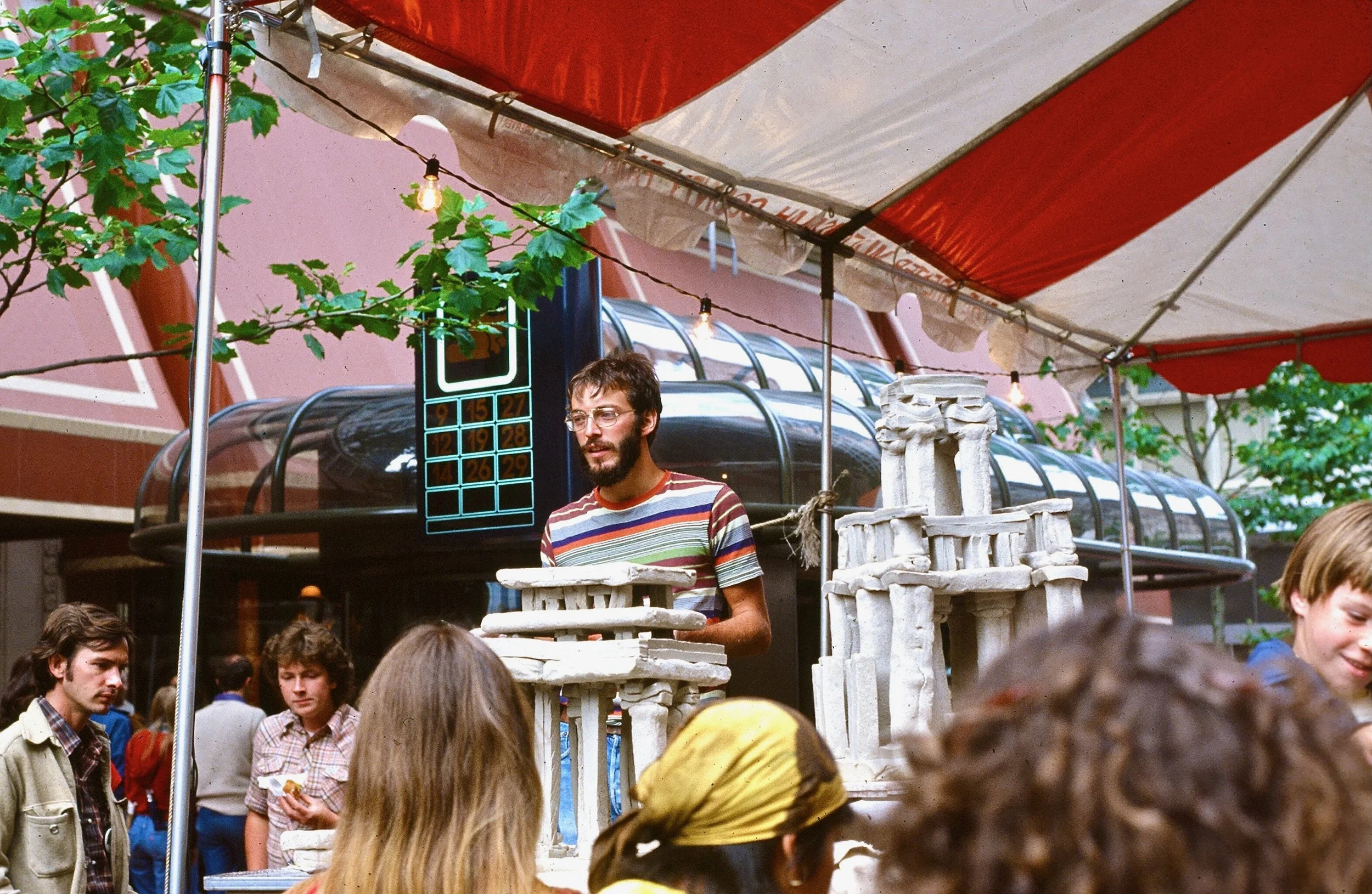 A man with glasses and a beard, wearing a striped shirt, stands behind a table of white plaster models at an outdoor market stall. There are several young people nearby, and a red and white striped canopy overhead. A large building and trees are in t