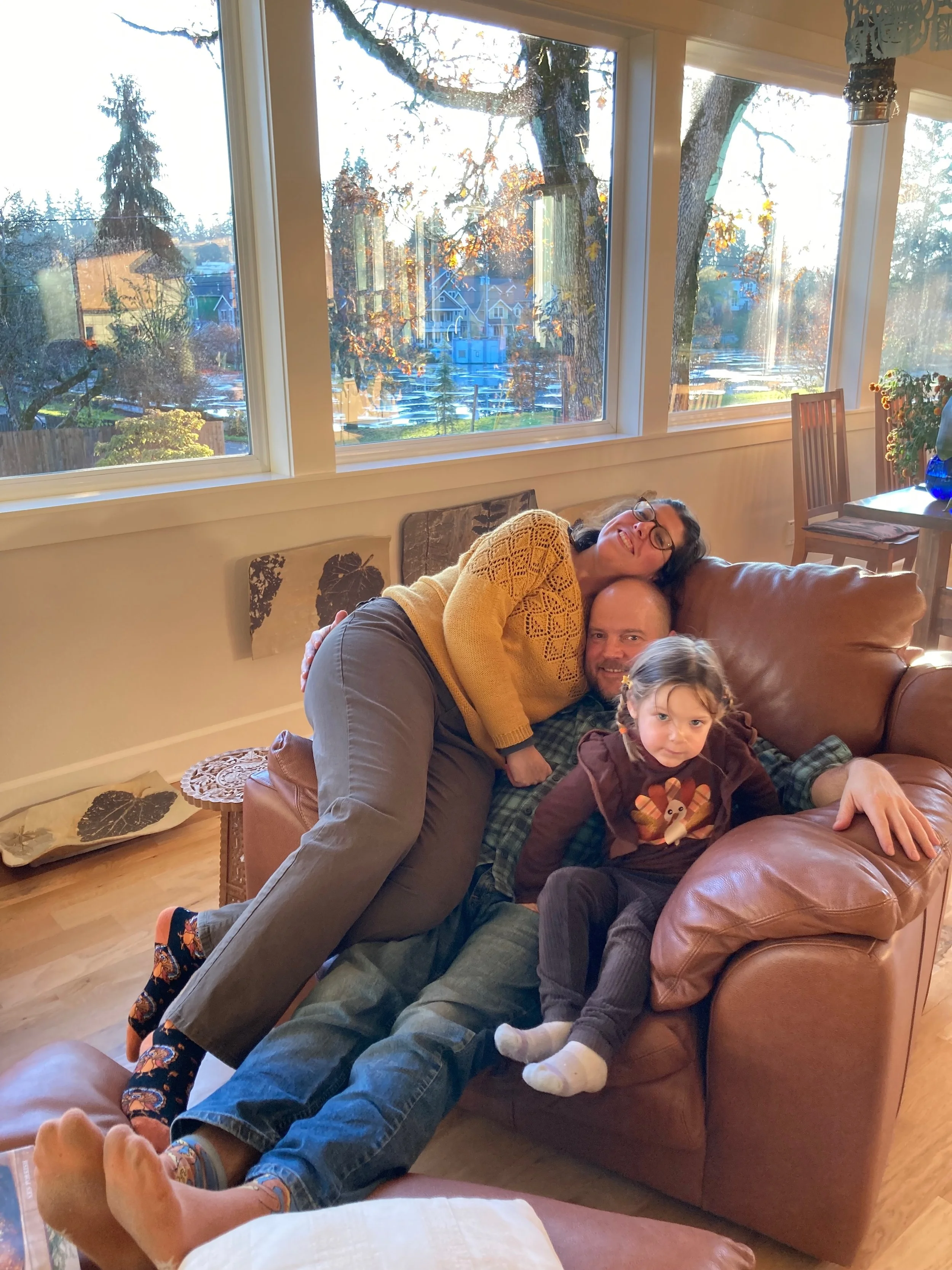 Family of three, two adults and a young girl, relaxing on a leather couch with a large window showing a scenic outdoor view of trees and houses in the background.
