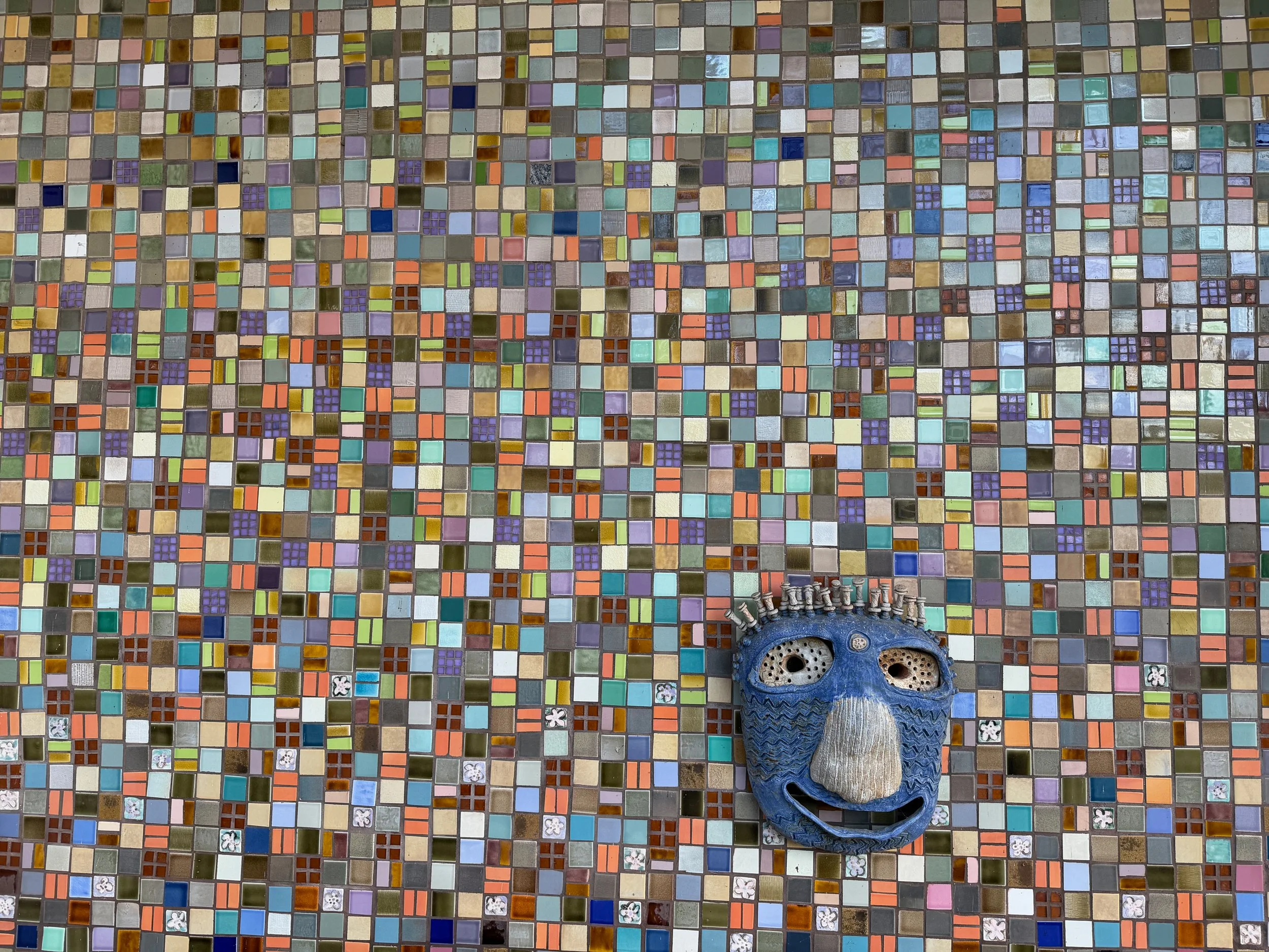 A decorative ceramic mask with a blue face, large nose, and wide smile, placed on a multicolored mosaic tile surface.