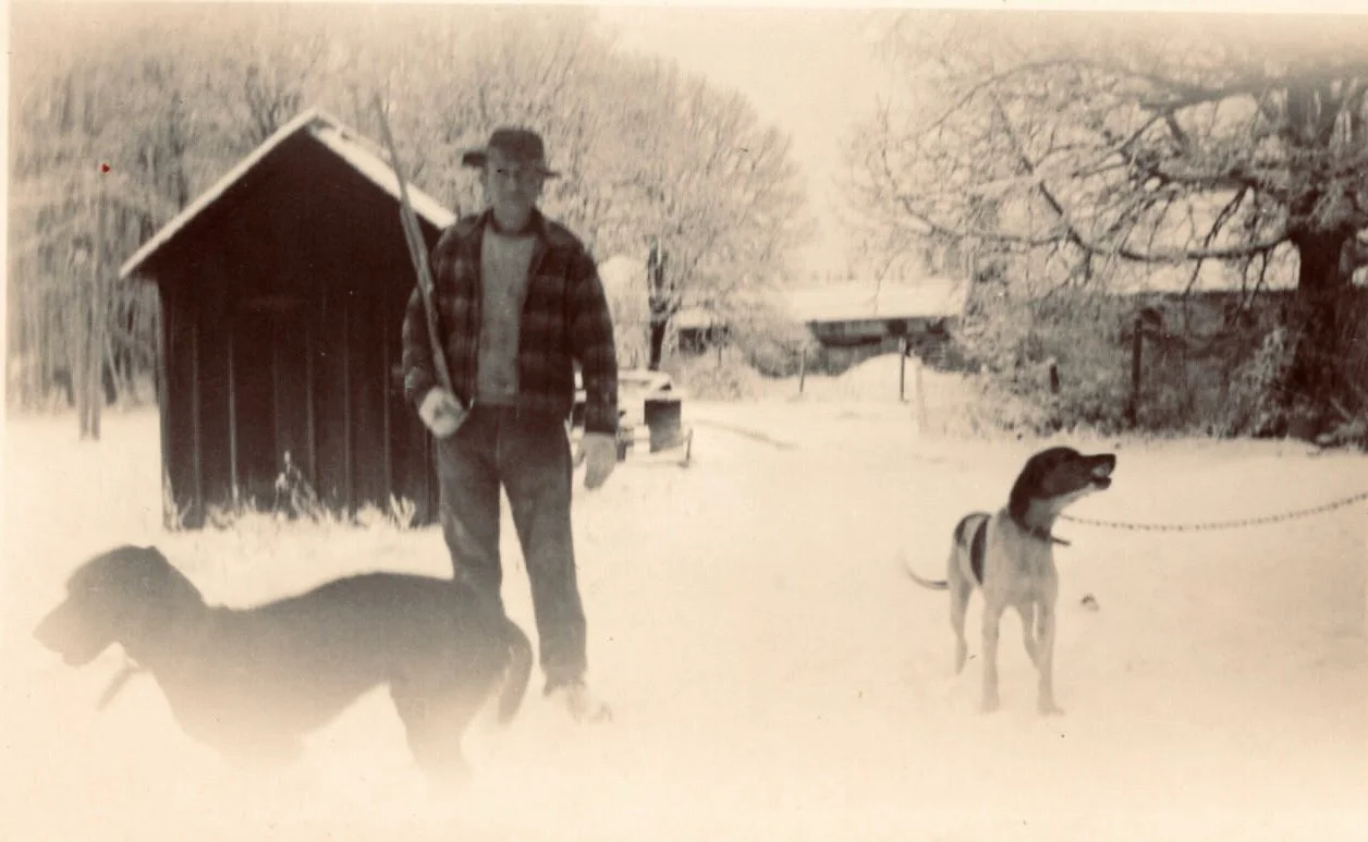 A man standing outdoors in the snow with two dogs, one on a leash, near a small shed and trees.