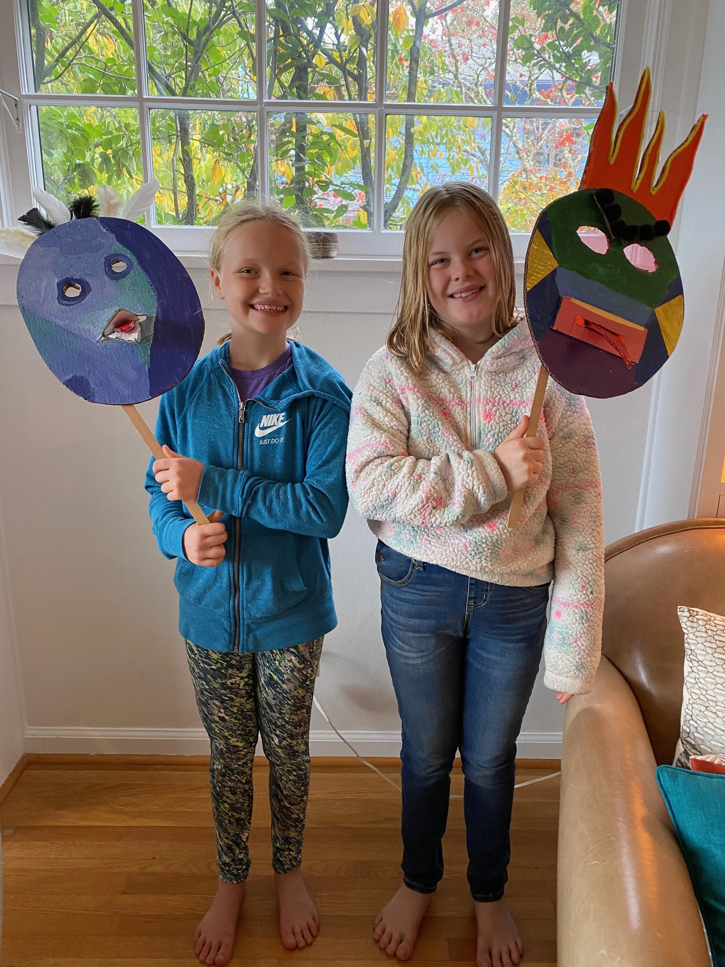 Two girls standing indoors, each holding a colorful paper puppet on a stick. The girl on the left is wearing a blue zip-up jacket and patterned leggings, smiling. The girl on the right is wearing a white fleece jacket and jeans, also smiling. Behind 