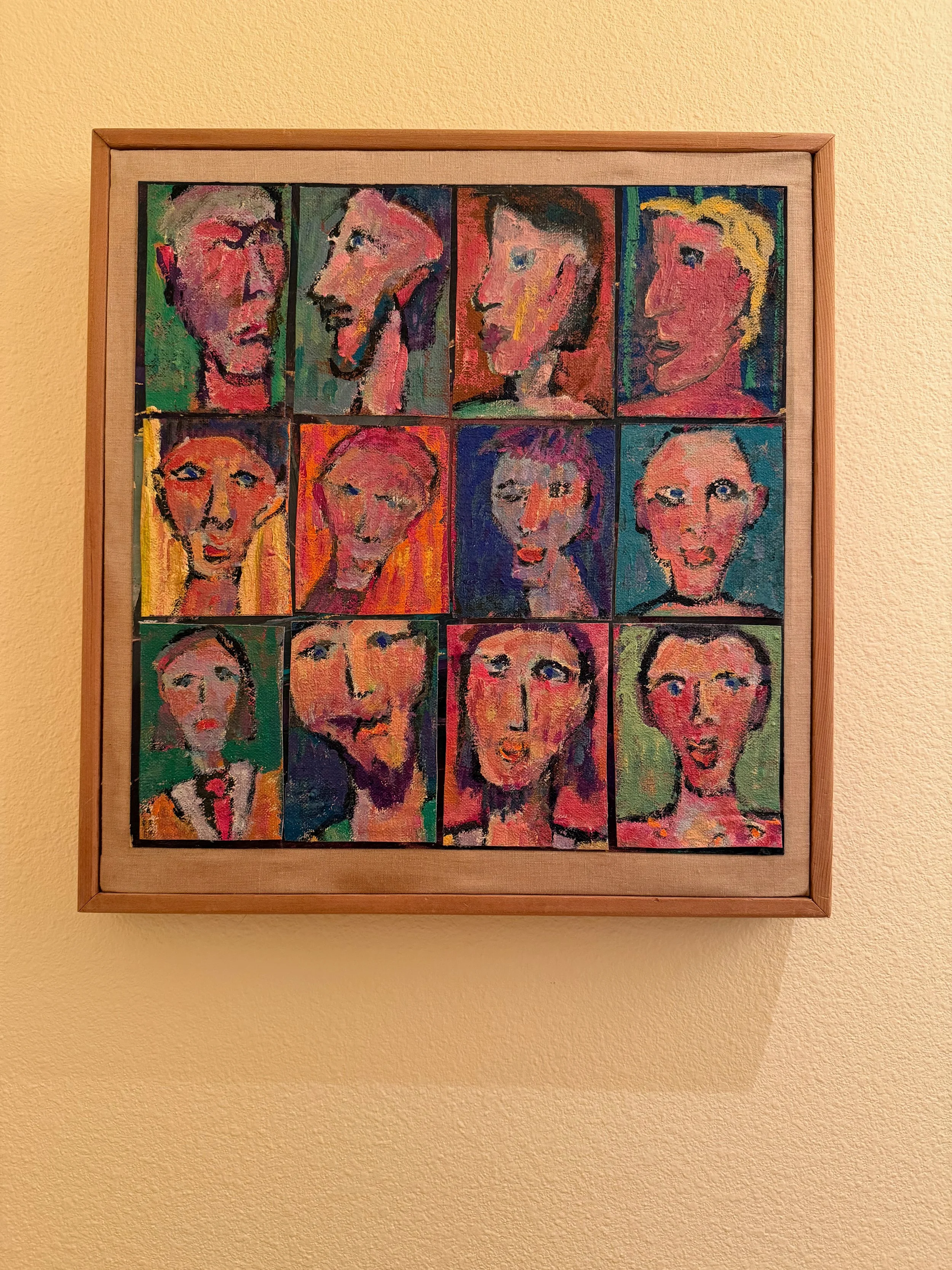 A framed colorful painting featuring twelve abstract portrait sketches arranged in a grid on a beige wall.