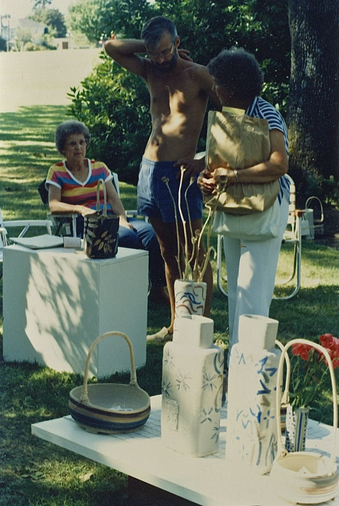 Three people outdoors at a gathering, with various pottery and decorations on tables in the foreground. An elderly woman is sitting, a shirtless man is standing, and a woman holding a bag is talking to him.