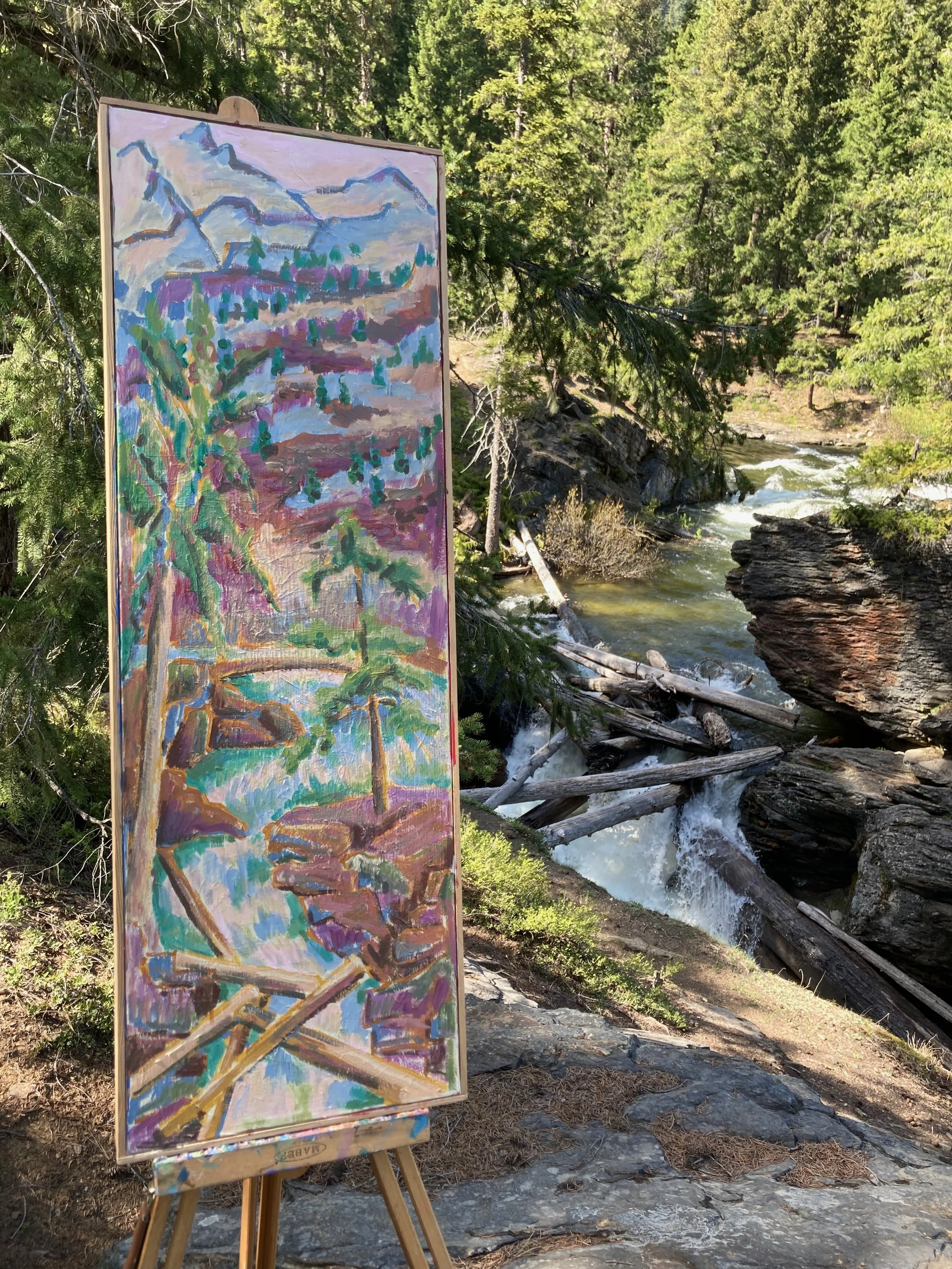 An outdoor painting of a mountain landscape with trees and a river on an easel in a forest next to a flowing creek.
