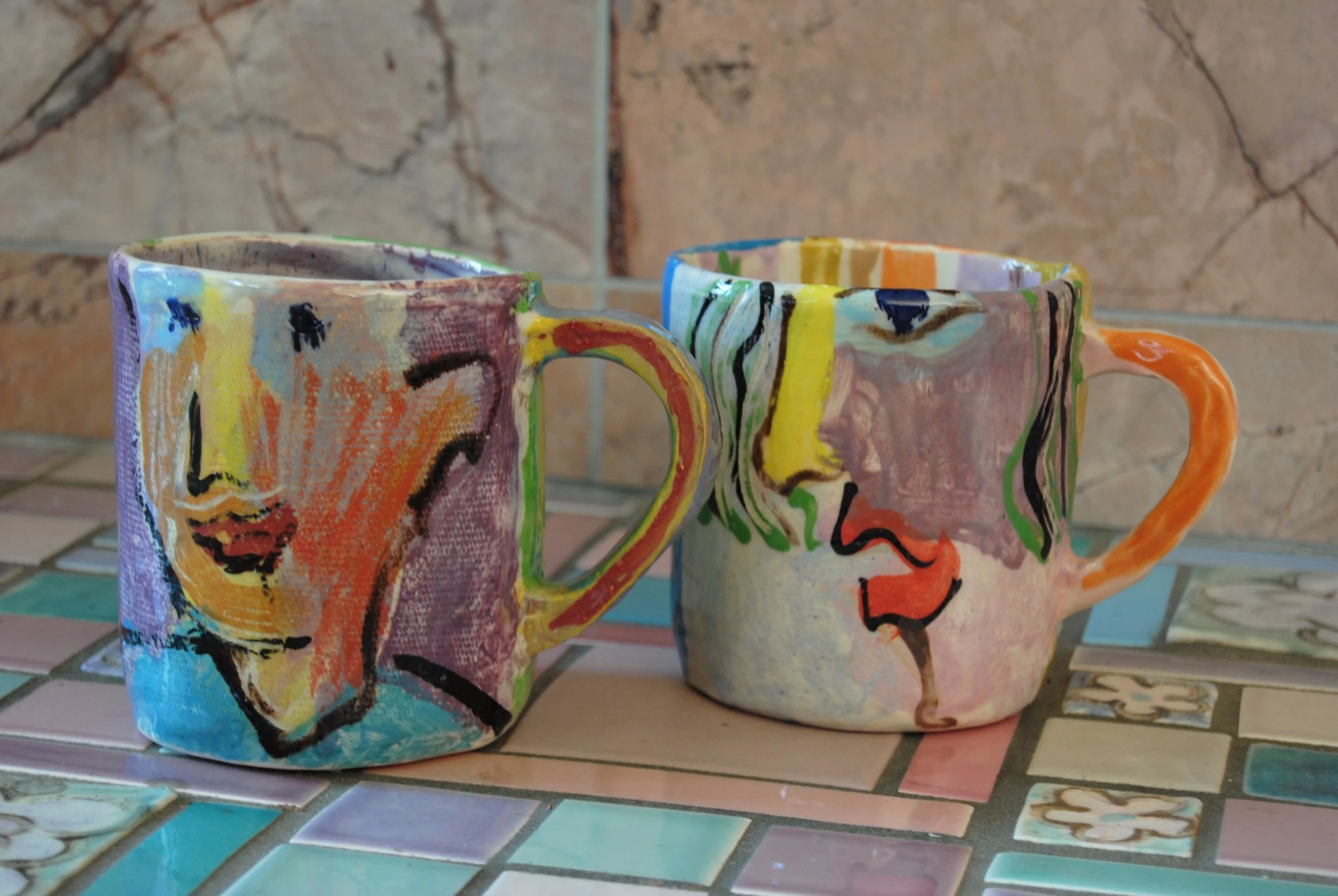 Two ceramic mugs with colorful abstract face designs sit on a tiled surface against a beige stone wall.