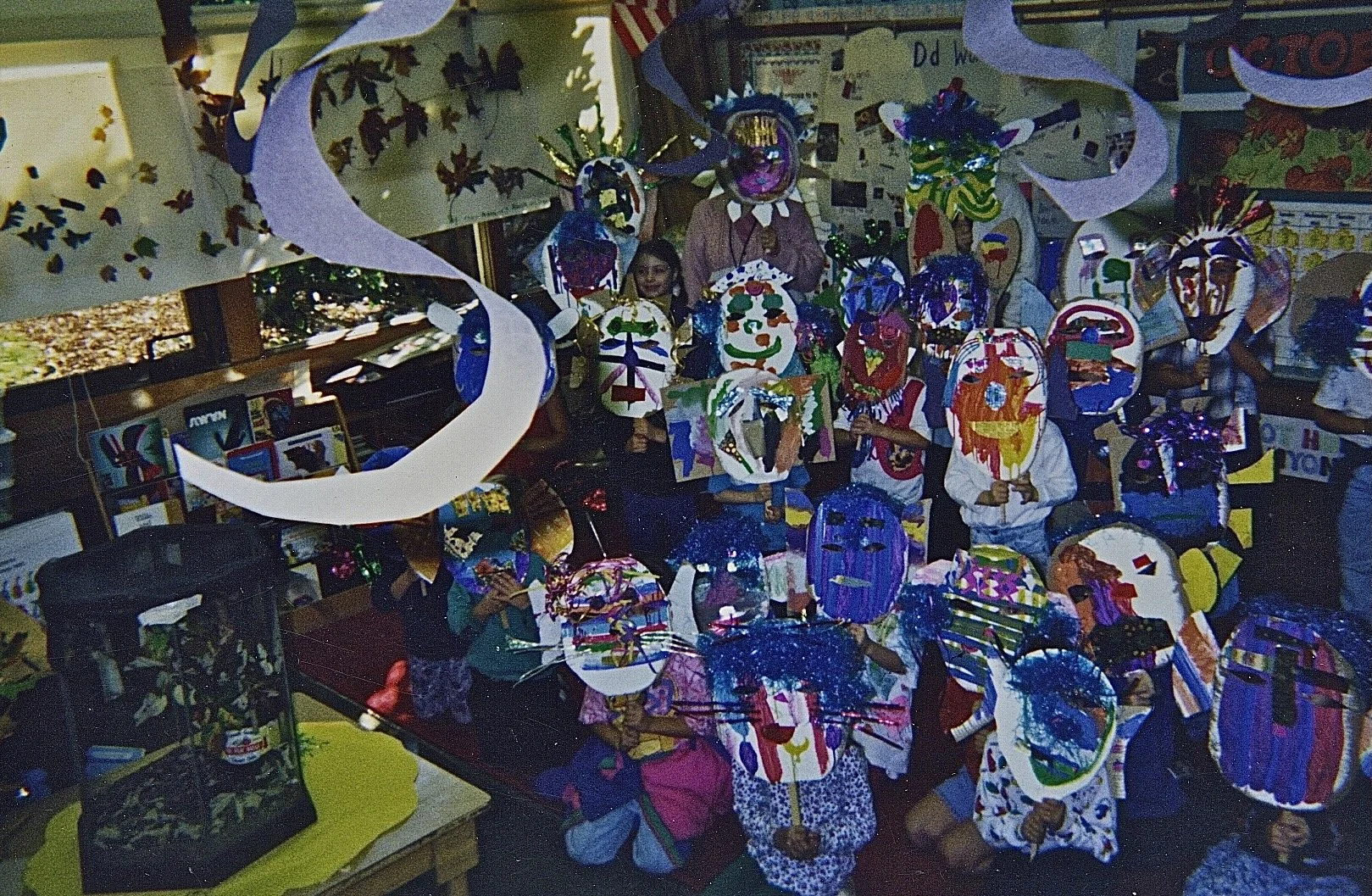Children in a classroom holding colorful paper masks and wearing Halloween costumes, with decorations hanging from the ceiling.