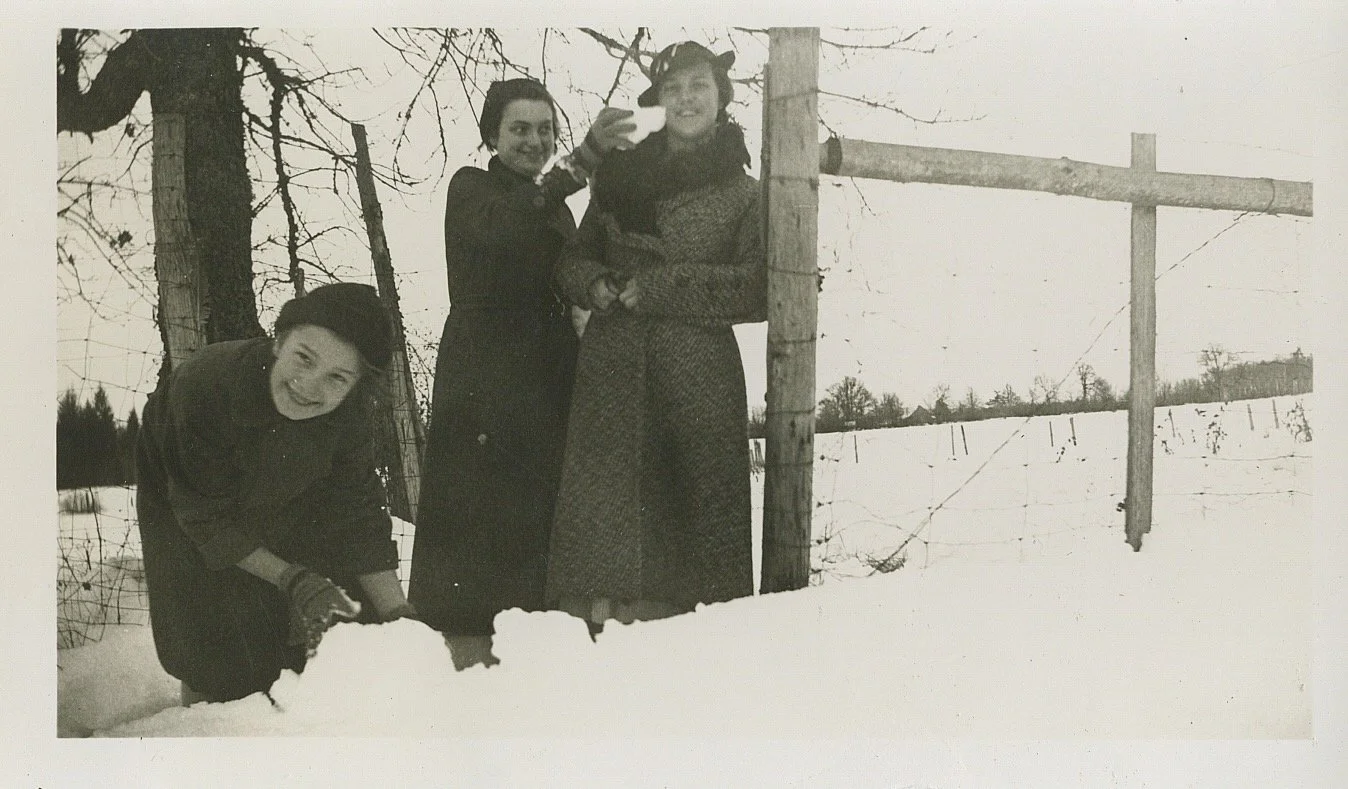 Three children outdoors in a snowy landscape, with a wooden fence and leafless trees in the background. Two of them are standing, one is bending down, all smiling, with one child taking a selfie.
