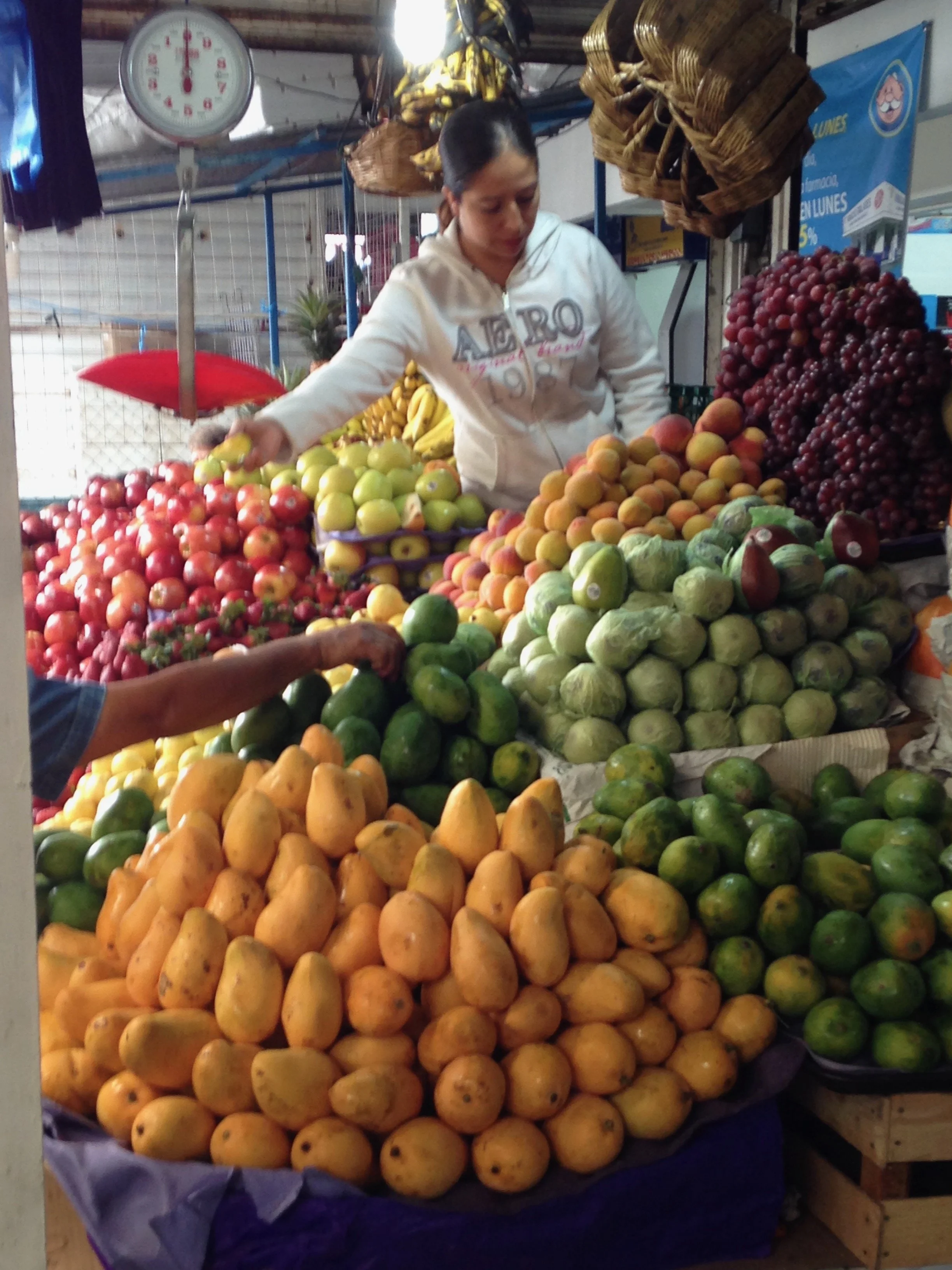 A woman at a fruit stand arranging various fruits including apples, peaches, limes, mangoes, grapes, and cabbages under a scale in an indoor market.