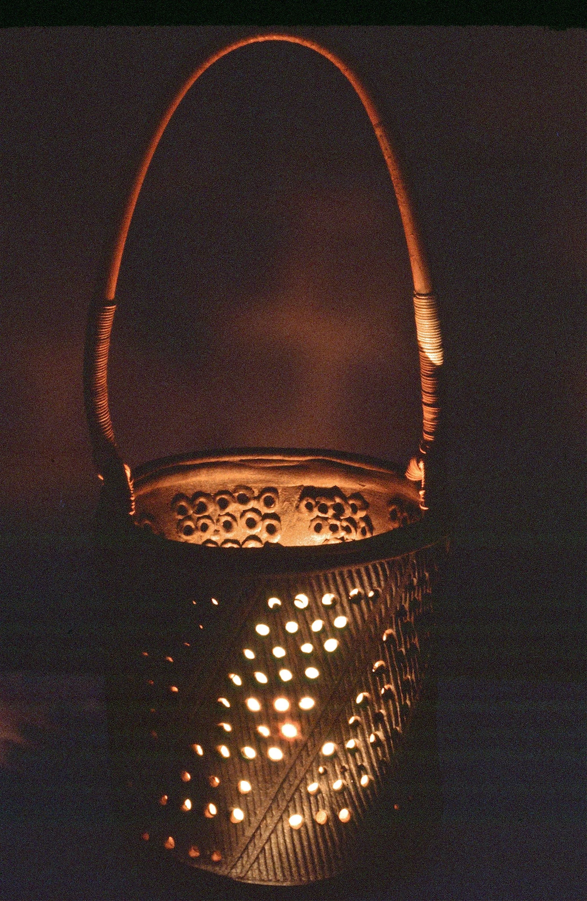 A glowing, perforated basket with a handle, illuminated from within in a dark setting.