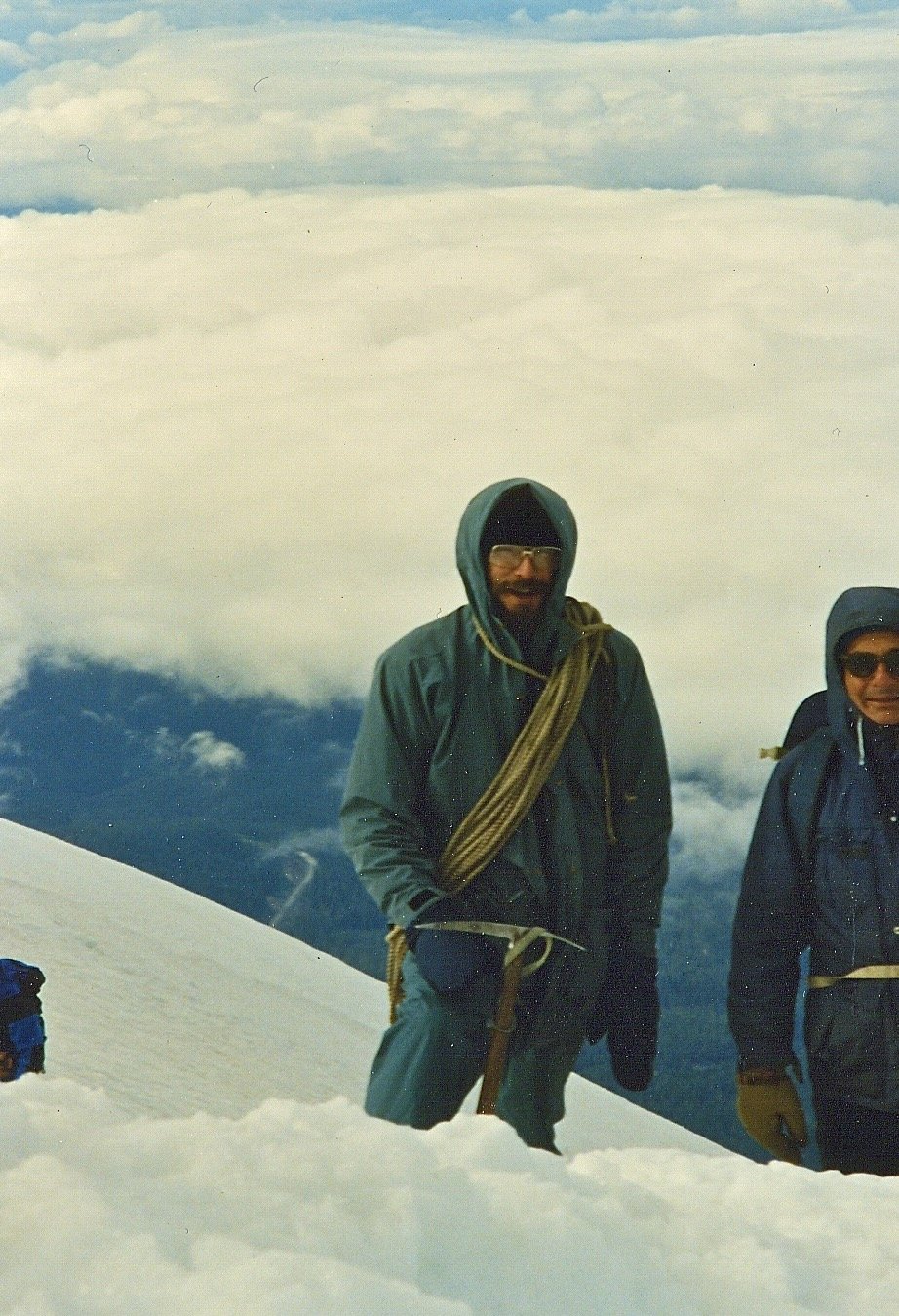 Two climbers in winter gear on a snowy mountain peak, with clouds and mountain scenery in the background.