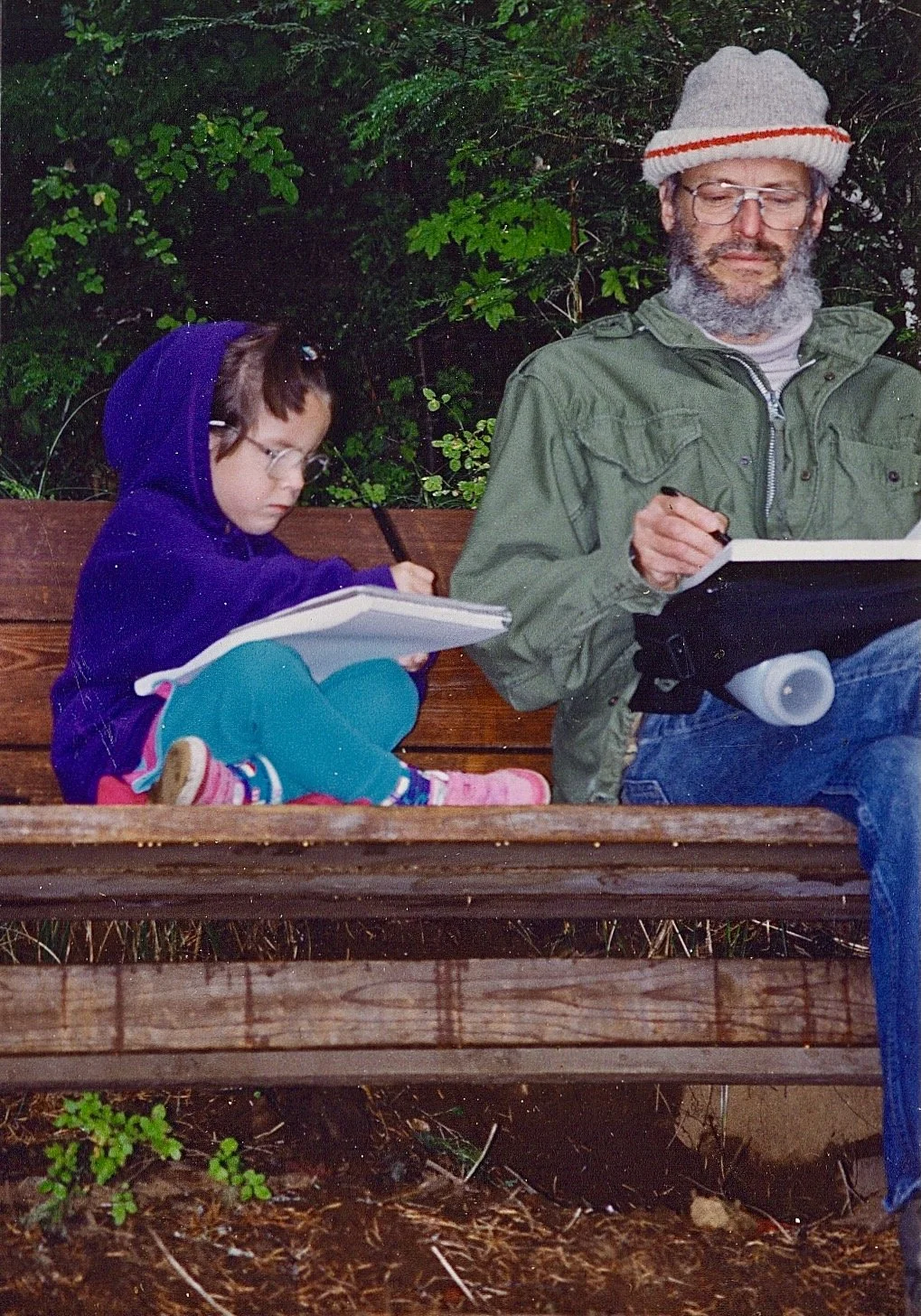 An adult man and a young girl sitting on a wooden bench outdoors, writing or drawing in notebooks, with green foliage in the background. The girl is wearing a purple hoodie and glasses, and the man is wearing glasses, a knitted hat, and a green jacke