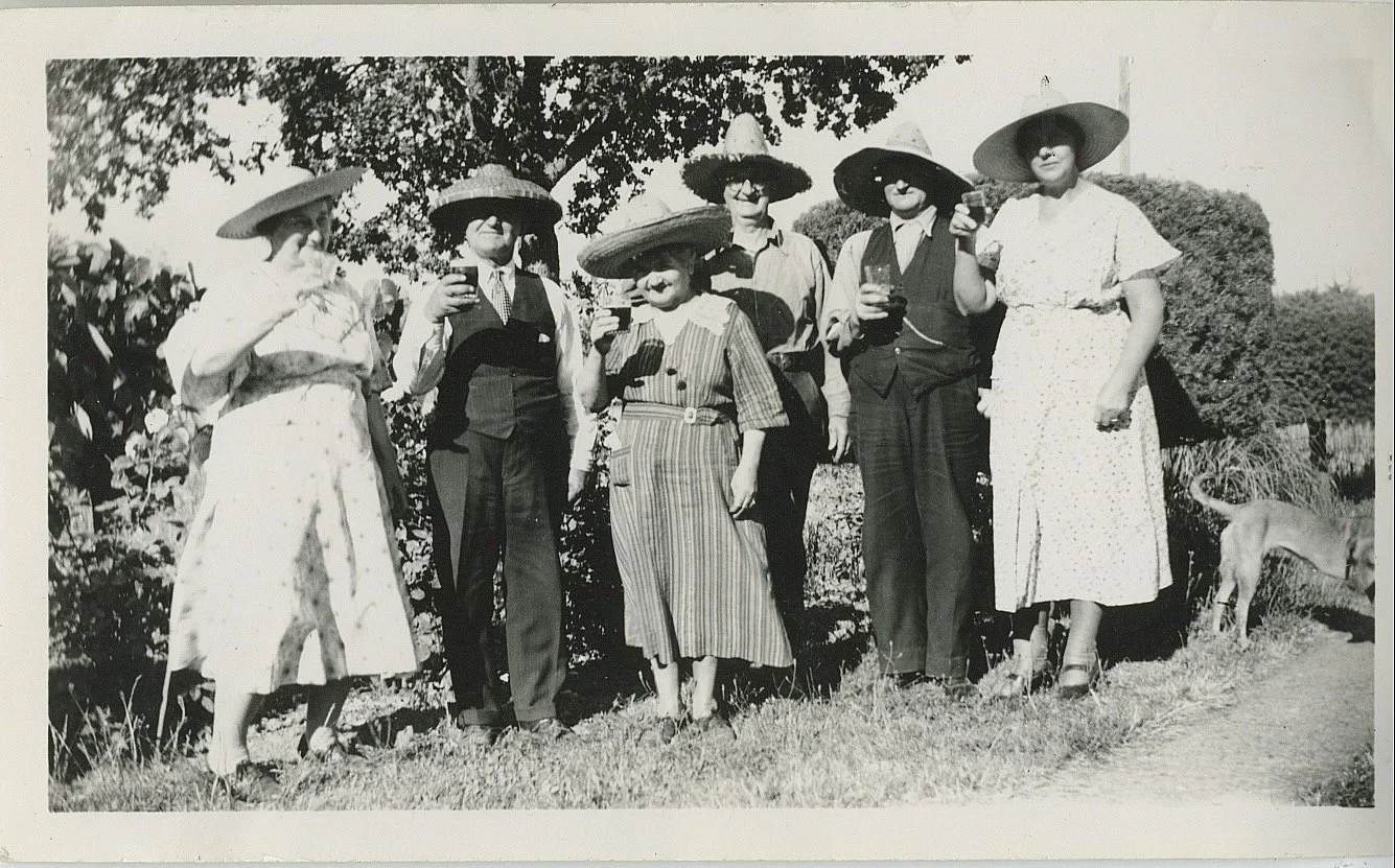 Black and white photo of six women in vintage clothing and large sun hats, standing outdoors on grass, some holding drinks, with a dog in the background.