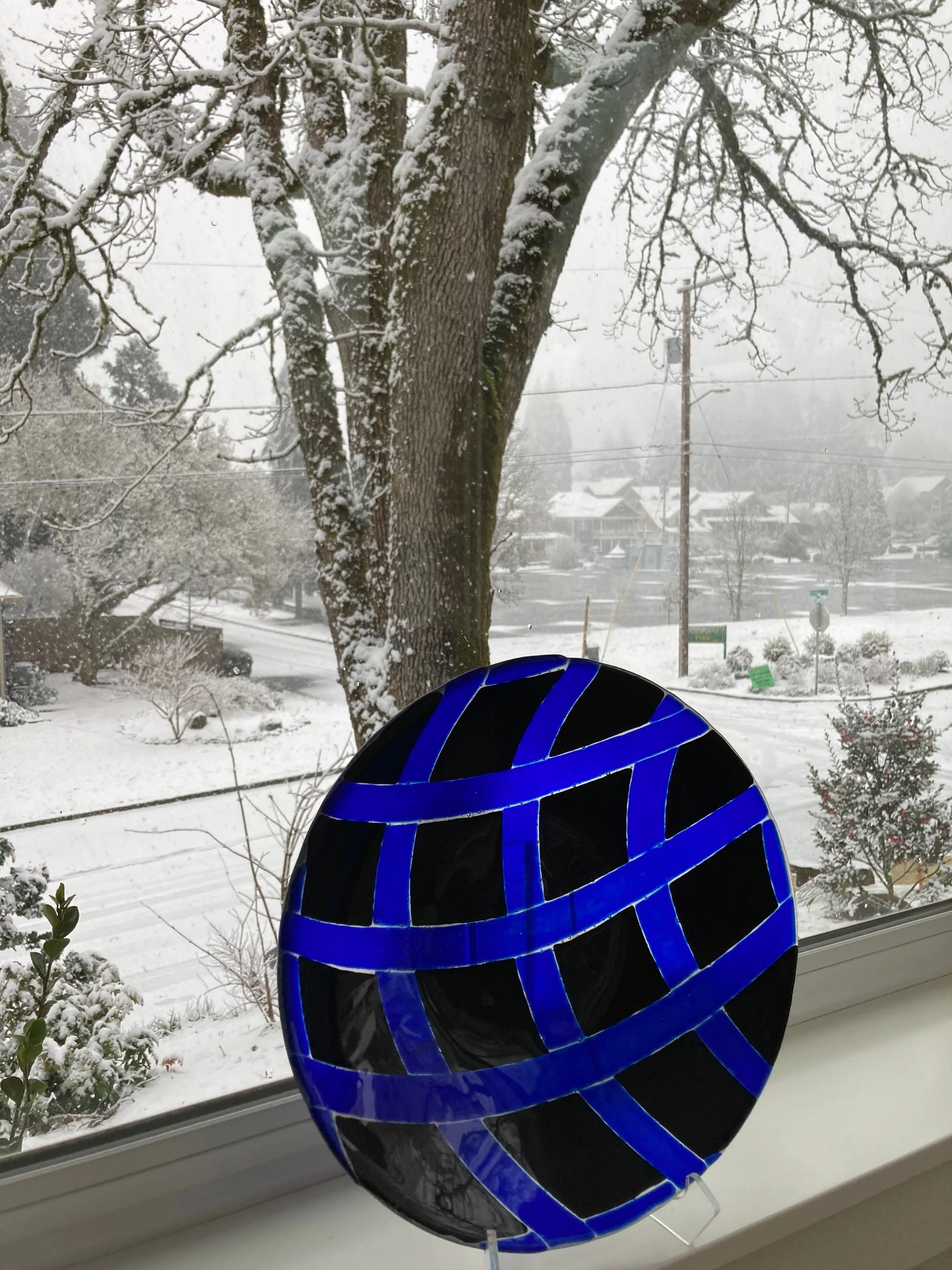 A blue and black decorative glass orb with a geometric pattern, placed on a window sill with a snowy outdoor scene visible through the window, including snow-covered trees and houses.