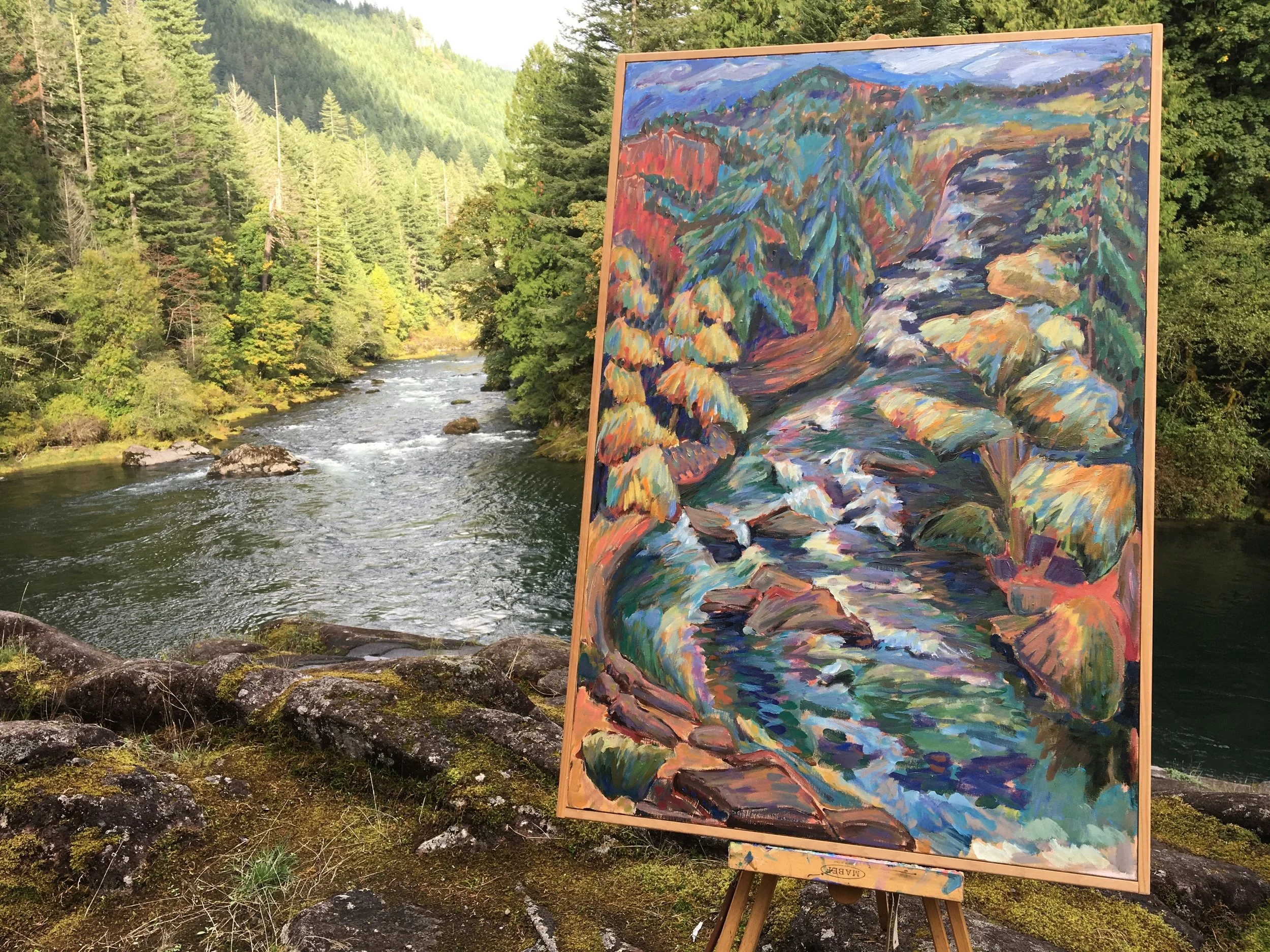 A landscape painting of a rushing river surrounded by trees with colorful foliage, set on an easel beside a river and lush forested mountains.