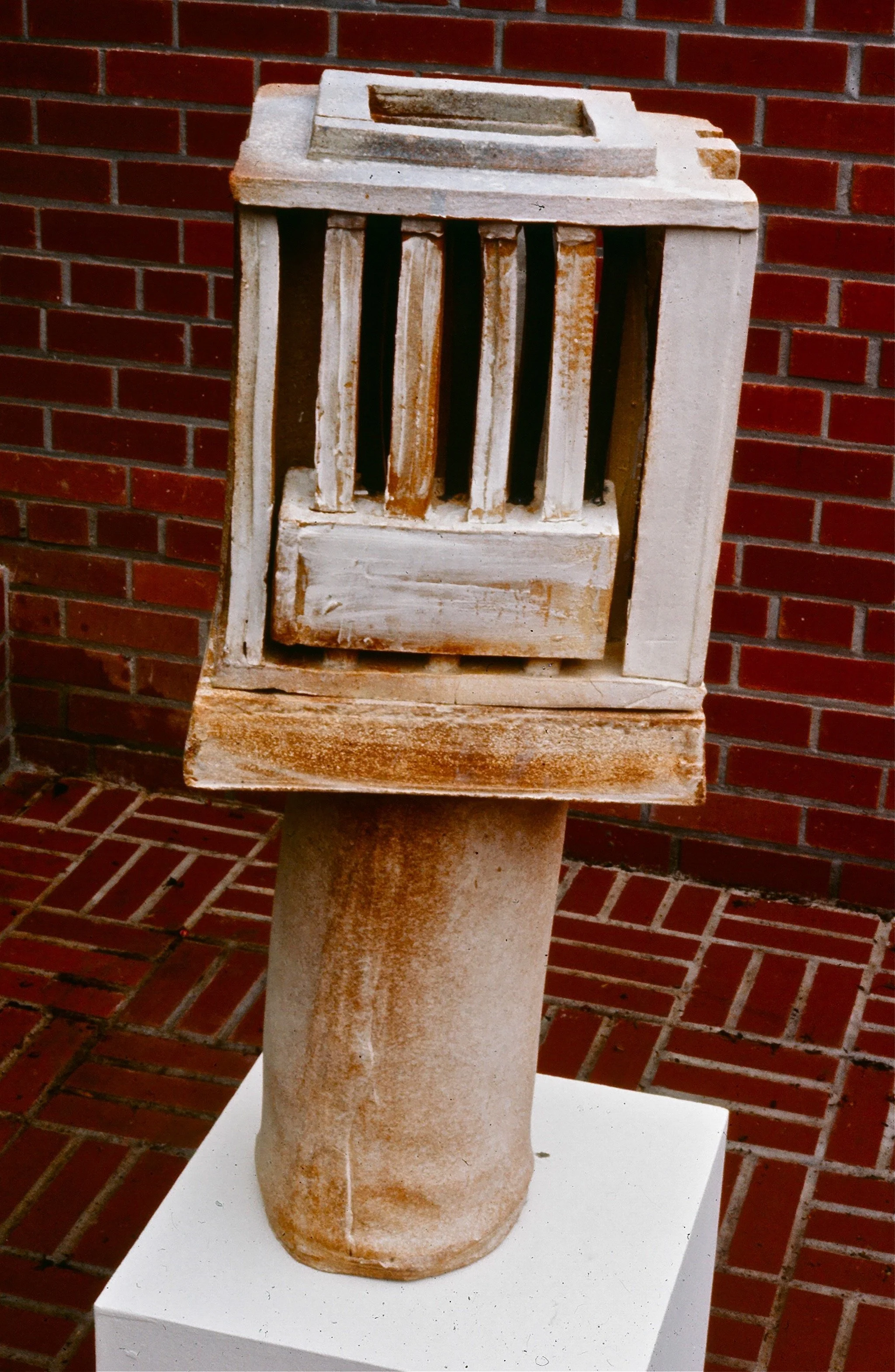 A stone sculpture featuring a rustic jail cell door with vertical bars, mounted on a round stone base, set against a red brick wall.