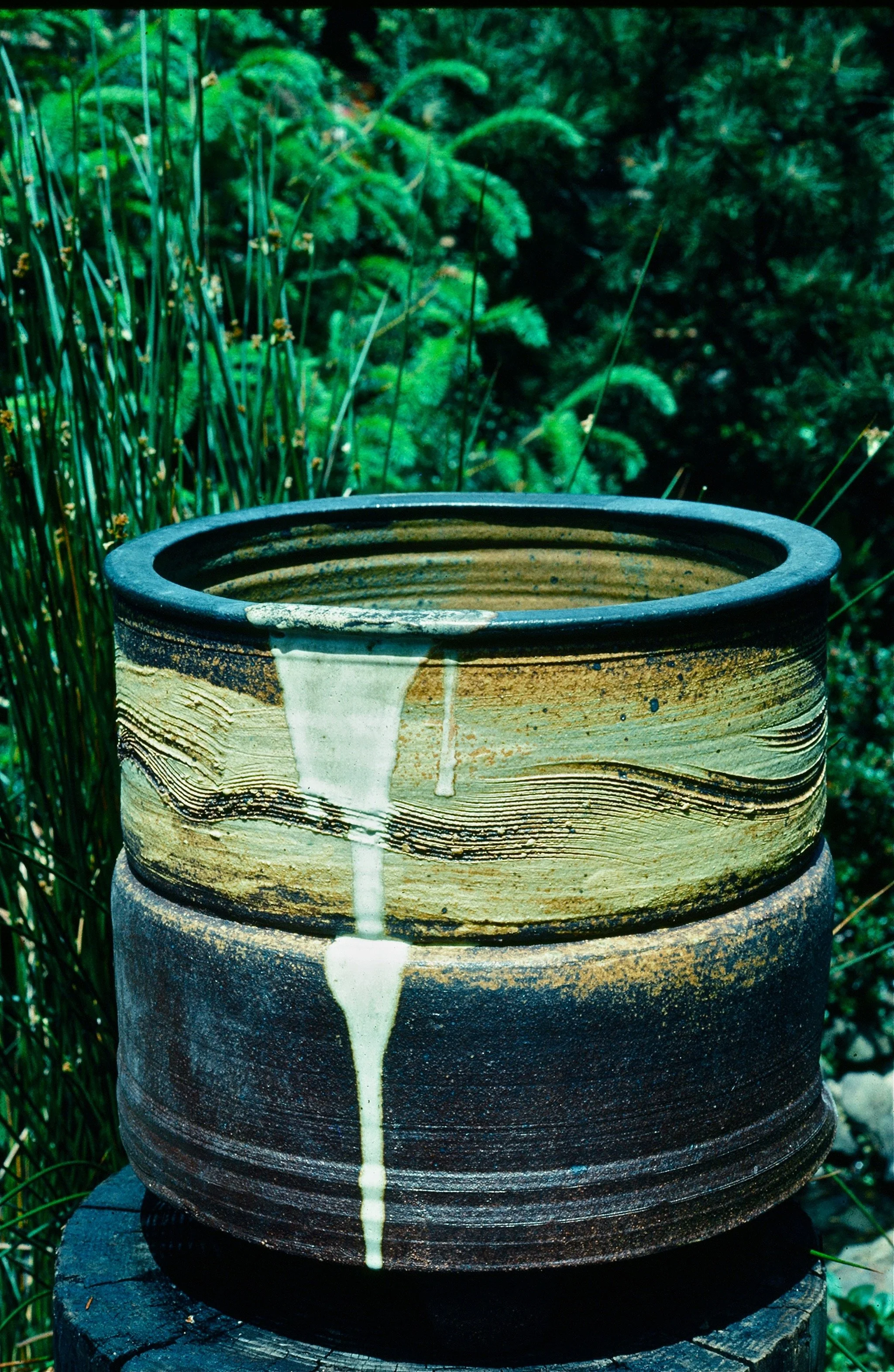 A ceramic pot with a brown and black textured glaze, sitting outdoors on a wooden stump with green plants and ferns in the background.