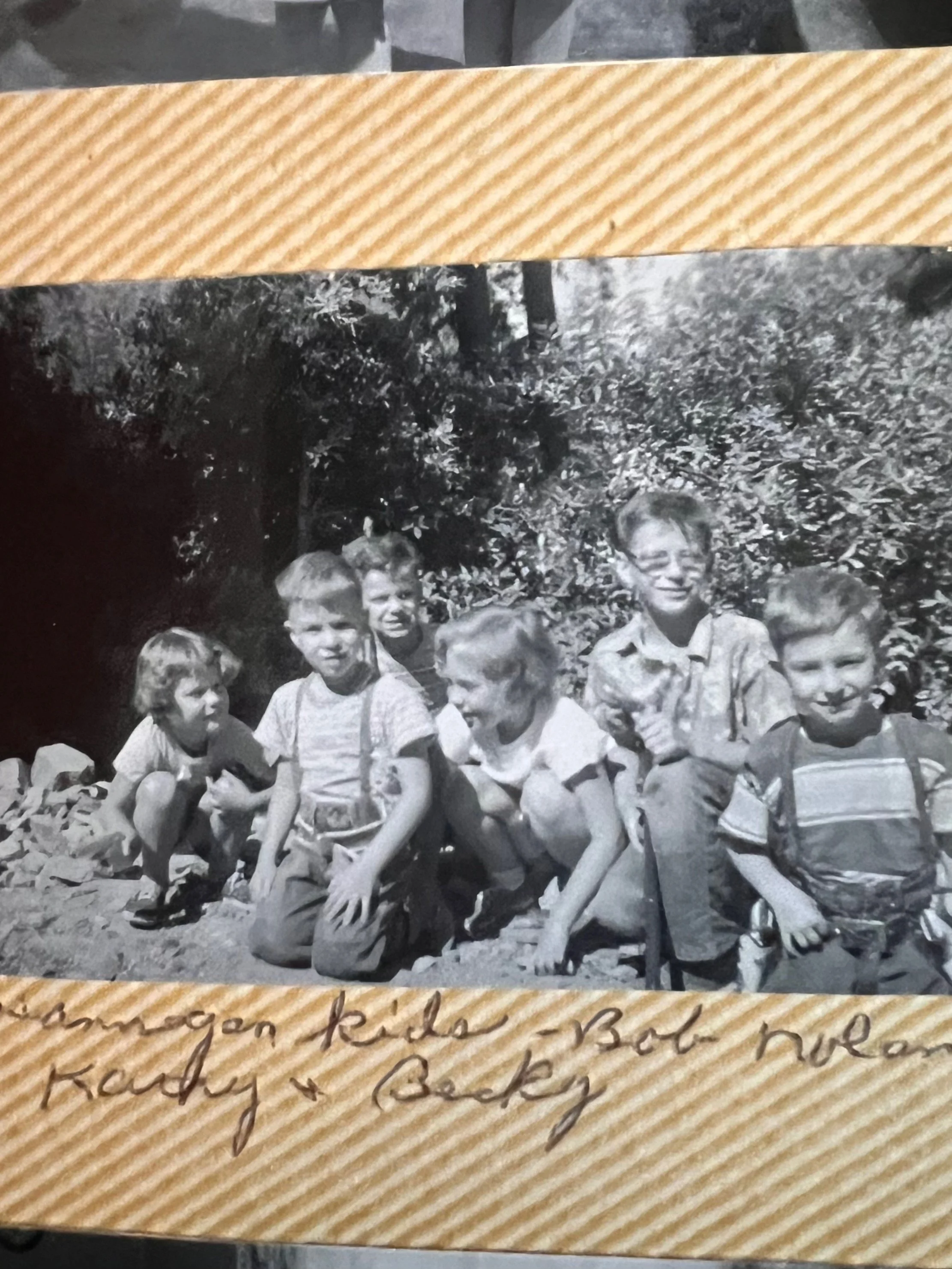 Black and white photograph of six children outdoors in front of bushes, crouching or kneeling on the ground, smiling and looking at the camera.