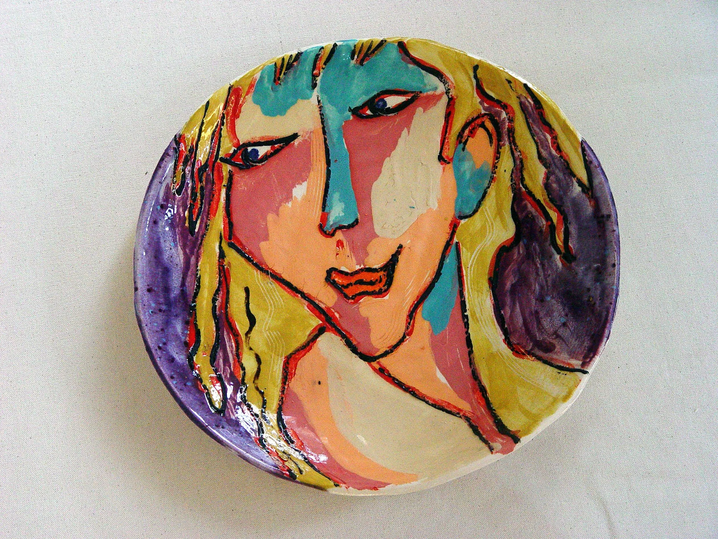 A decorative ceramic plate with a colorful abstract portrait of a woman with long yellow hair, blue eyes, and red lips.