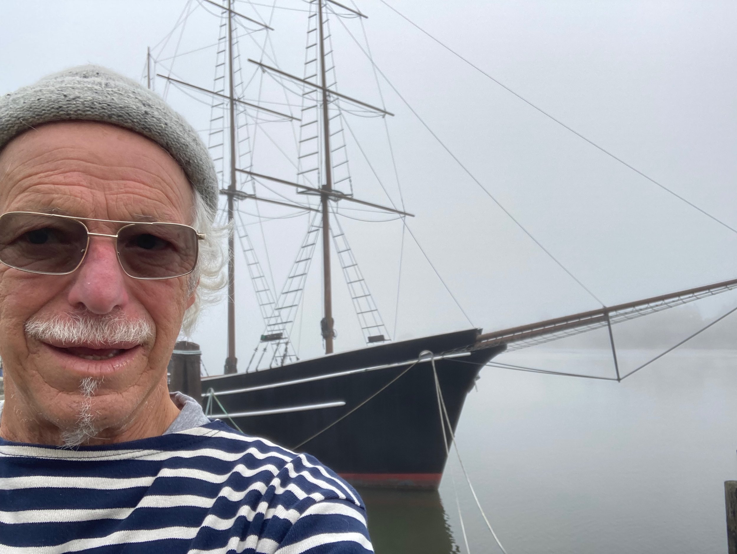 An elderly man with white hair and glasses taking a selfie in front of a tall, historic sailing ship on a foggy day.