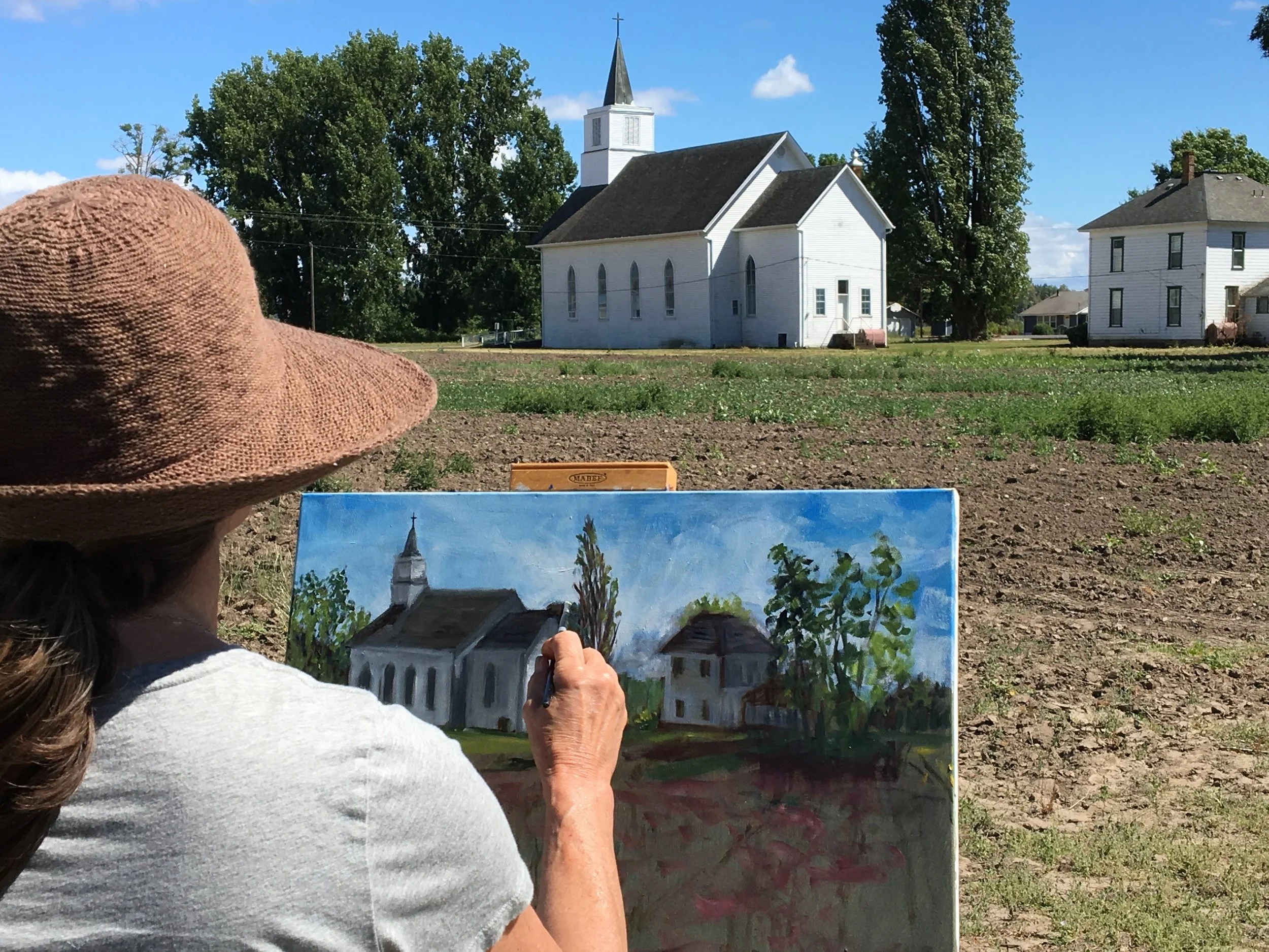 Woman with a wide-brimmed hat painting a church scene on canvas outdoors while looking at the real church in the distance.