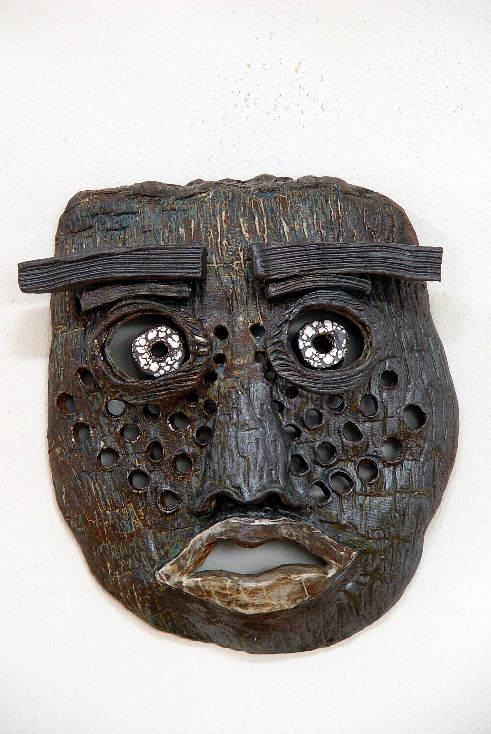 A wooden mask with a dark finish, featuring circular holes for eyes and mouth, with prominent eyebrows, and carved details on a white wall background.
