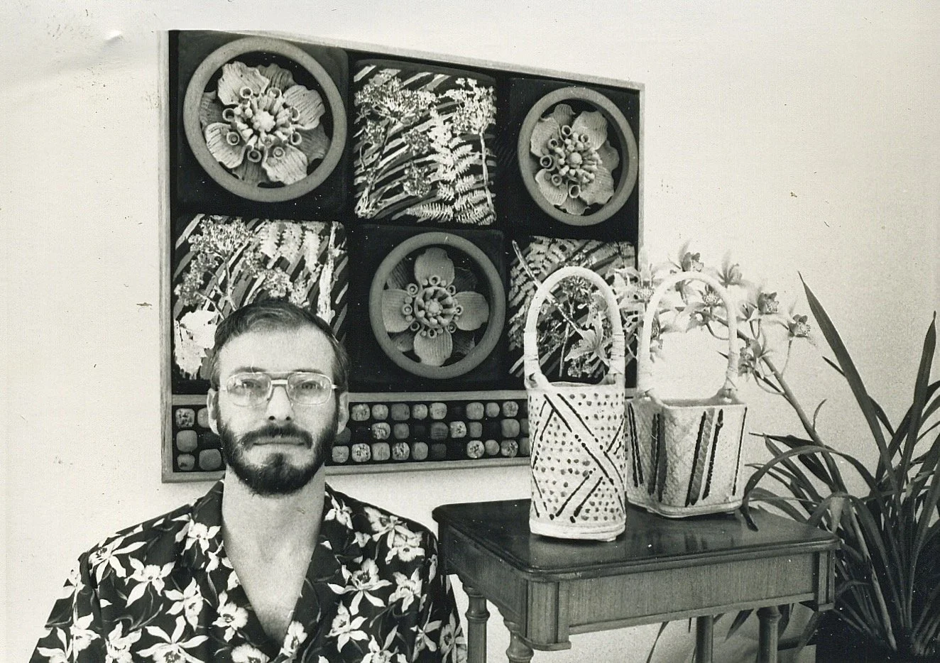 A man with glasses and a beard wearing a floral shirt sitting in front of a table with potted plants and a colorful art piece on the wall behind him.