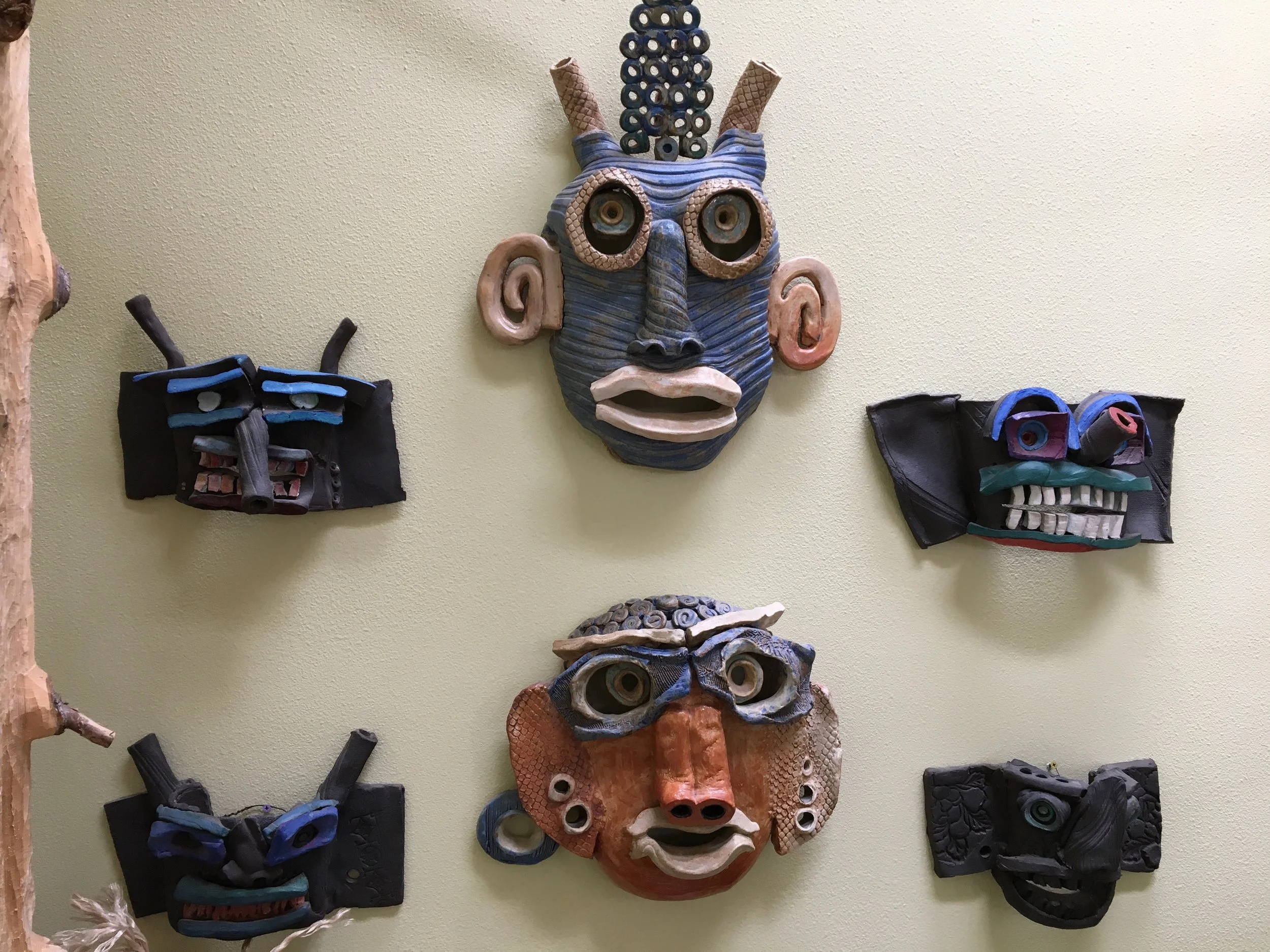 Collection of colorful, handmade masks on a yellow wall, featuring various expressive faces with exaggerated features and decorative elements.