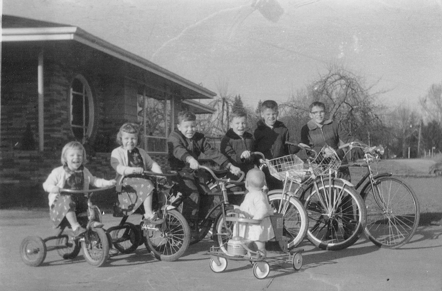 Black and white photo of seven children smiling outdoors, some sitting on bicycles and a tricycle, with two standing next to their bikes, in front of a house with a sloped roof and a leafless tree in the background.