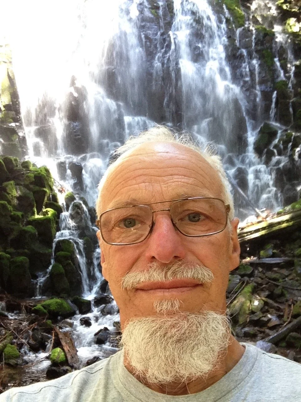 An elderly man with glasses, white hair, and a white beard taking a selfie in front of a waterfall.