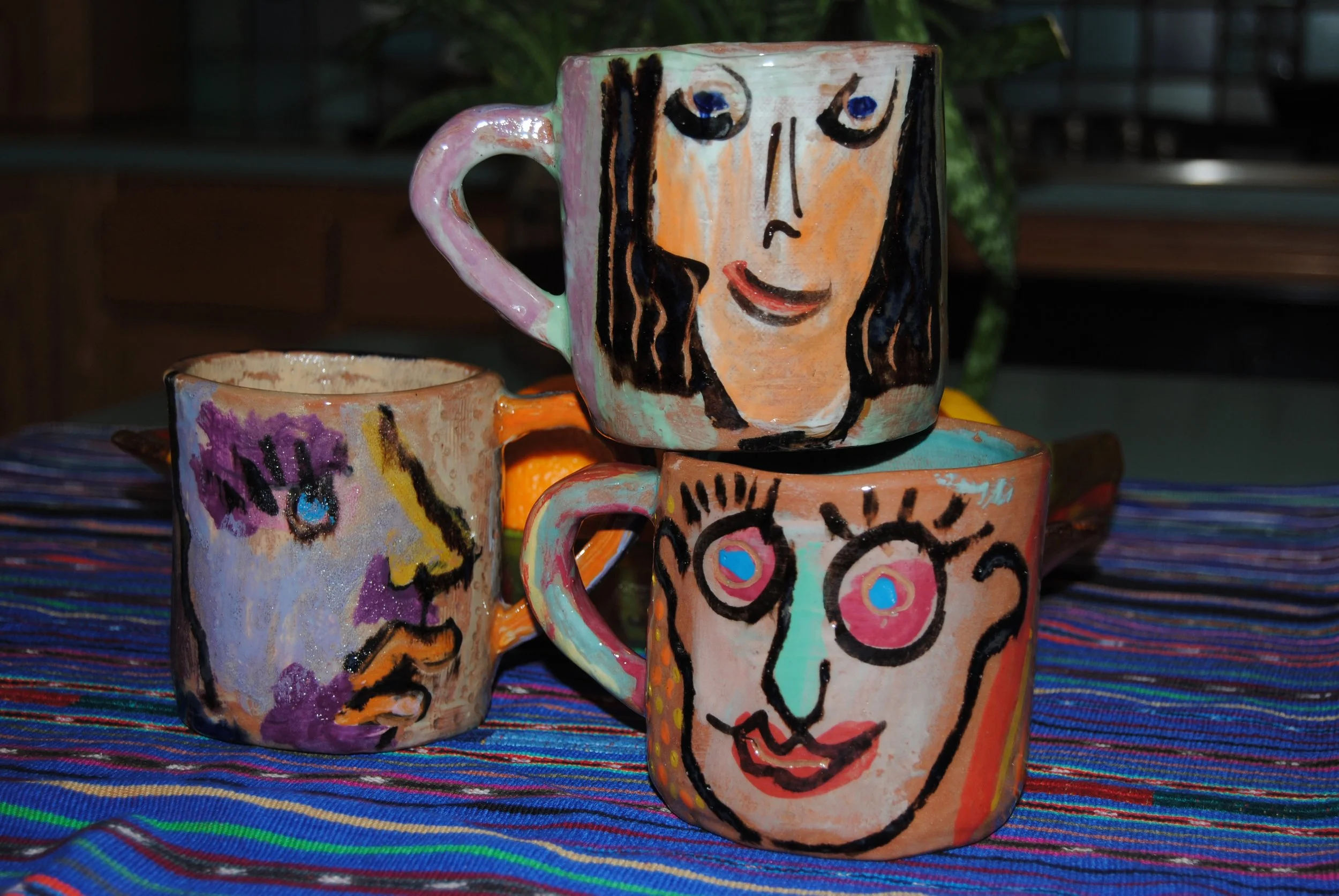 Three ceramic mugs with abstract face designs, placed on a colorful striped cloth.