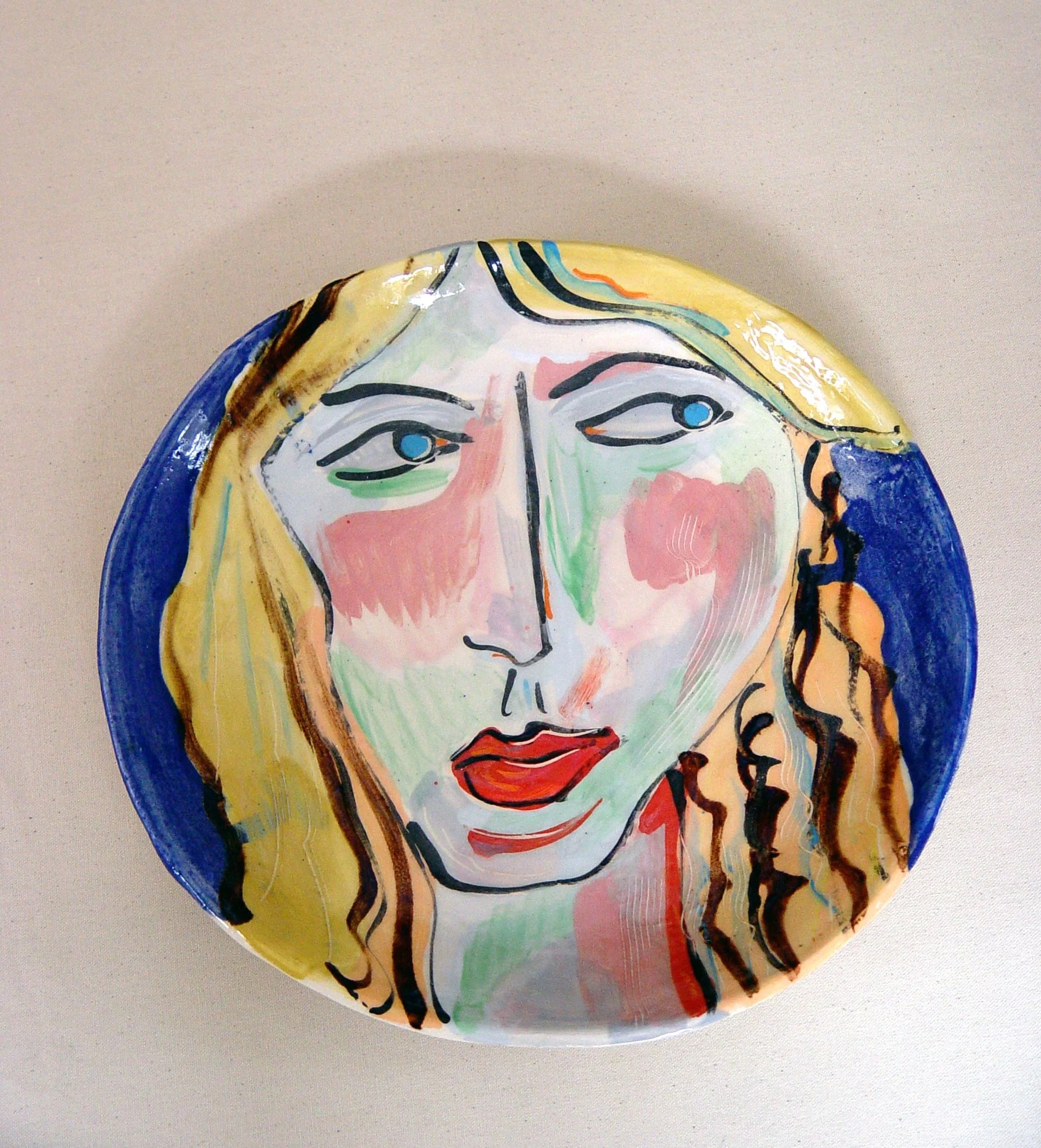 Hand-painted ceramic plate featuring an abstract portrait of a woman's face with large eyes, red lips, and long hair, using bold brushstrokes and vibrant colors.