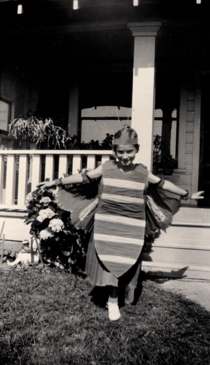 A young girl dressed in a homemade butterfly costume with wings, standing on a sidewalk in front of a house with a porch and flowers, smiling with arms outstretched.