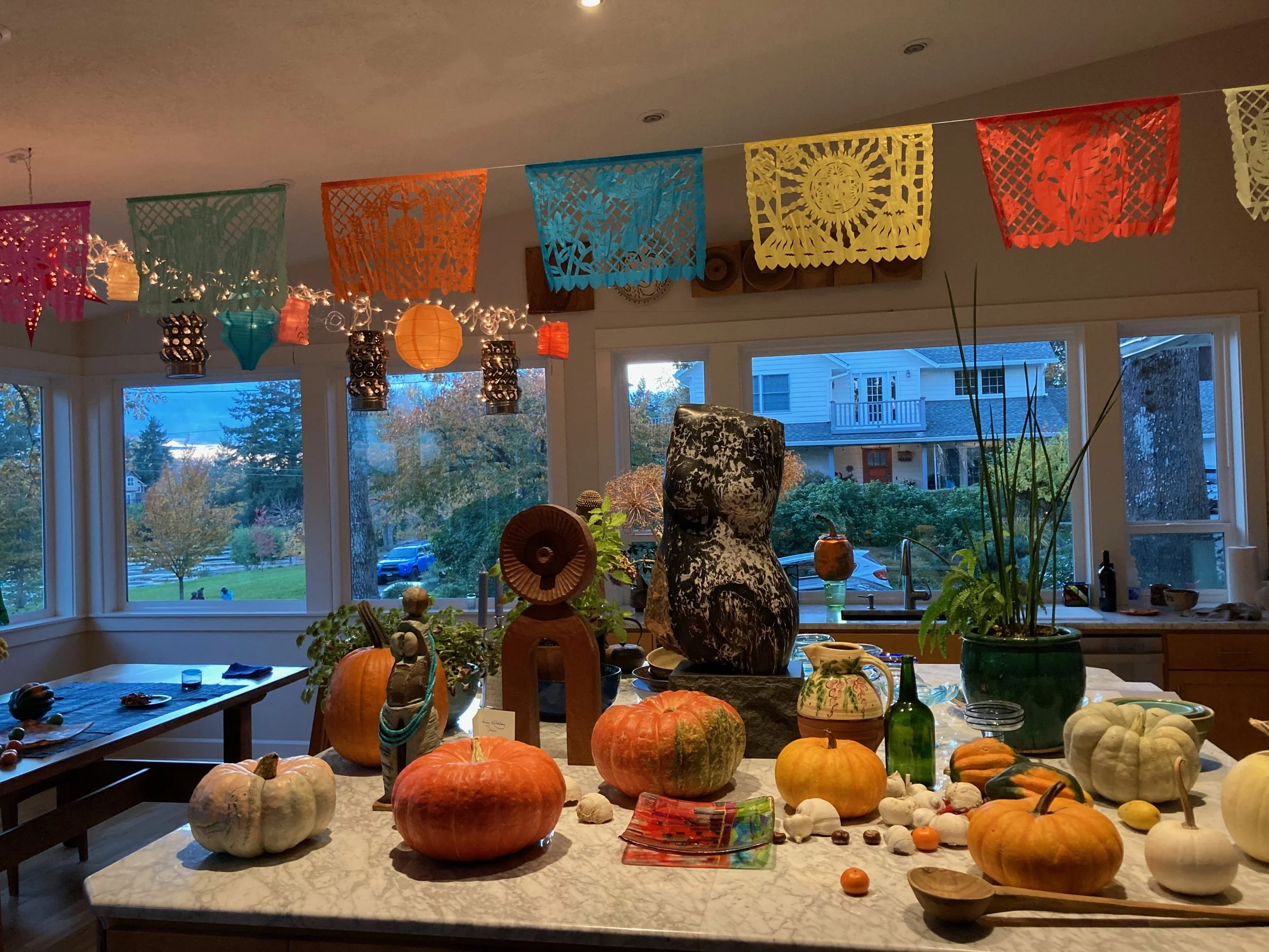 An indoor room decorated for fall or Día de los Muertos with colorful papel picado banners, paper lanterns, and various decorative pumpkins and gourds on a kitchen island, with large windows showing trees and houses outside.