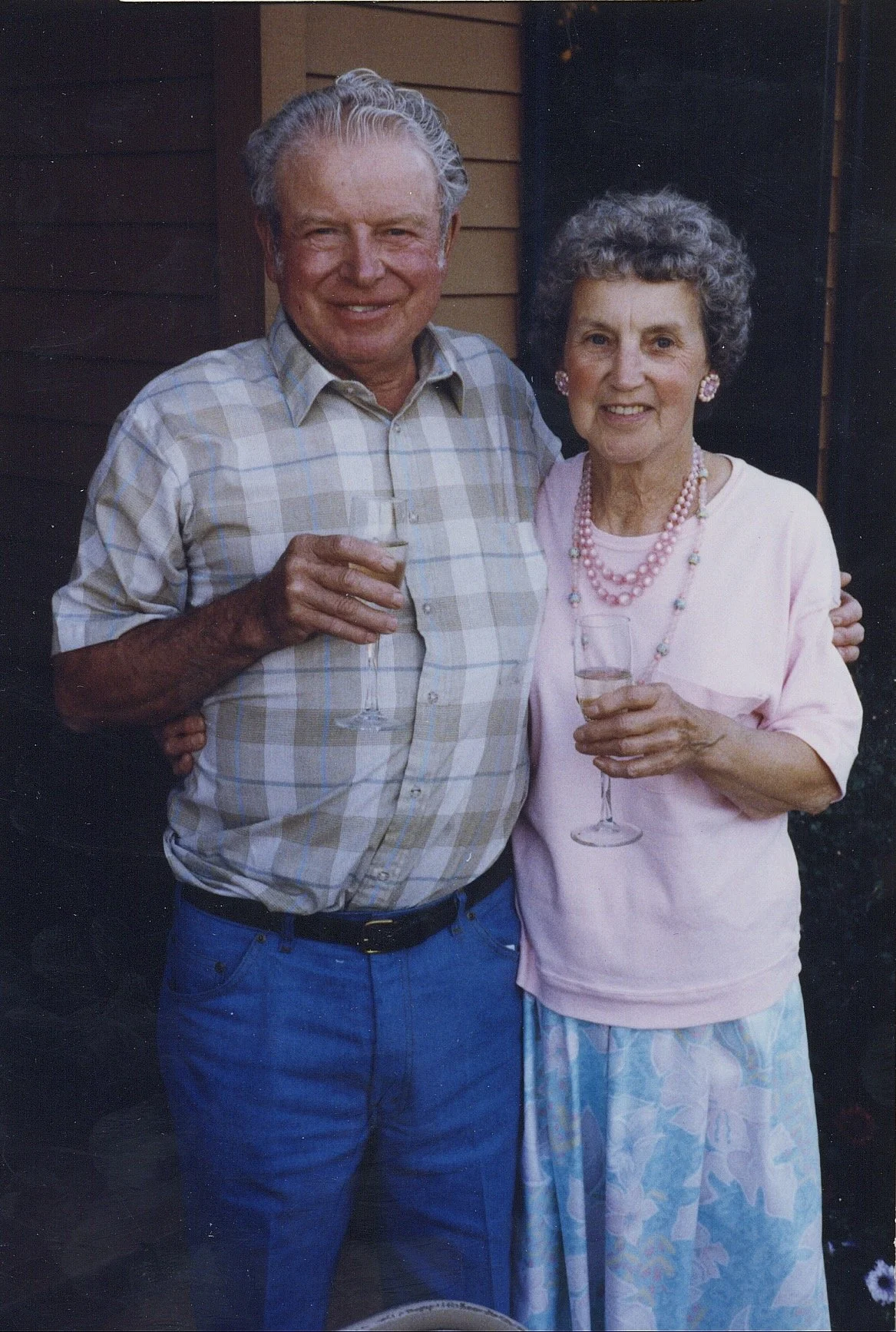 An elderly man and woman smiling, standing close together, each holding a glass of champagne. They are dressed casually, with the man wearing a light plaid shirt and jeans, and the woman wearing a light pink top, a floral skirt, and pink jewelry. The