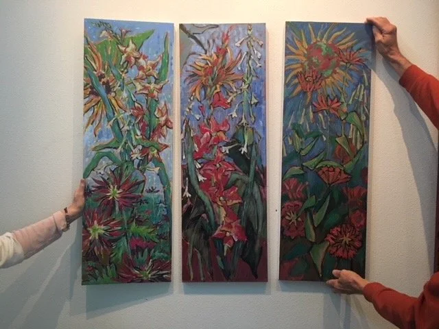 Three vertical abstract floral paintings with vibrant colors, held by two people against a plain wall.