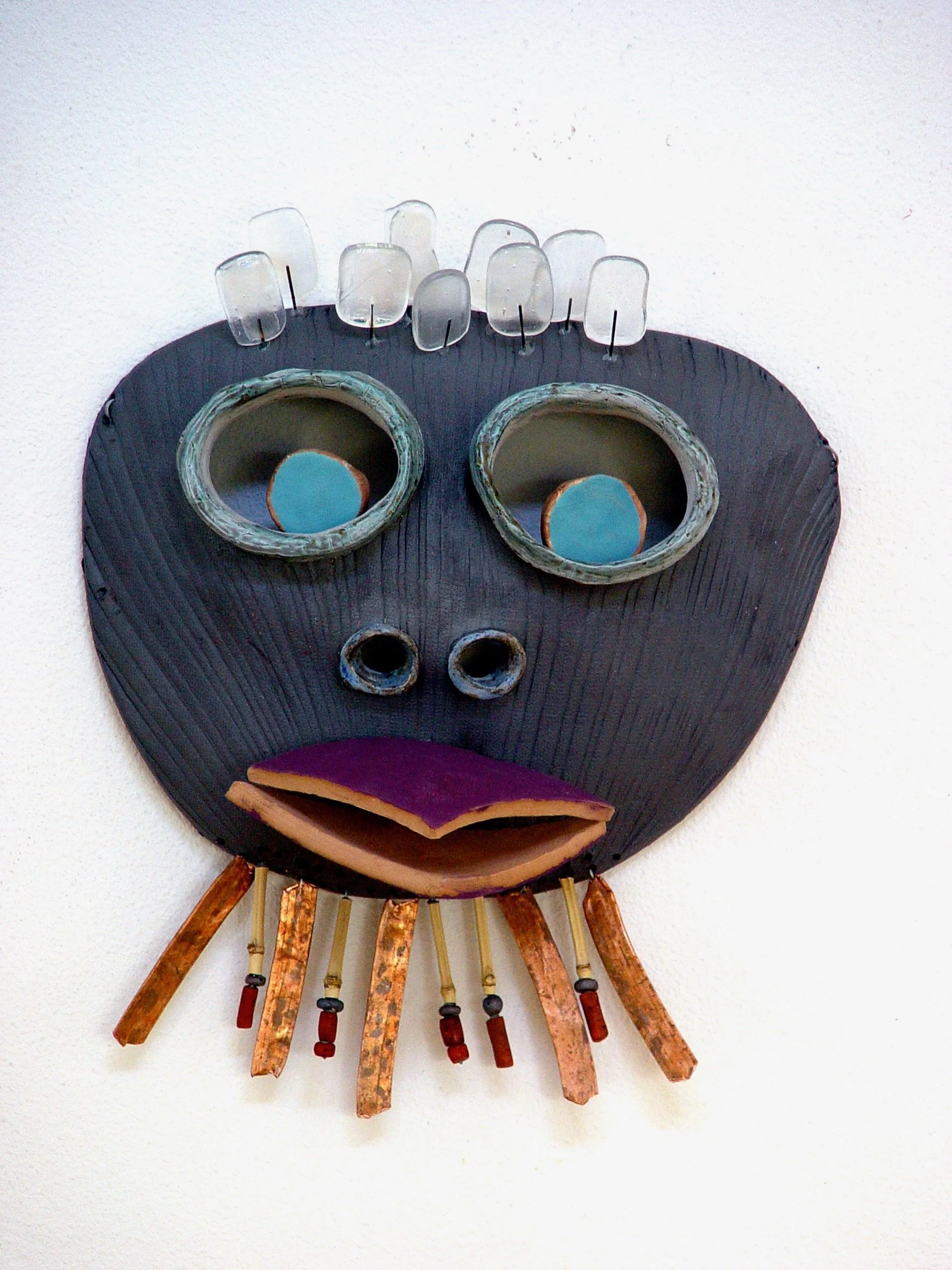 An artistic mask resembling a face, made of various materials including black wood or ceramic for the face, glass for the eyes, and other mixed media elements, mounted on a white wall.