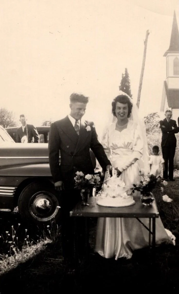 A black and white photo of a bride and groom at their wedding outdoors. The groom is wearing a suit with a boutonniere, and the bride is in a wedding dress with long sleeves. They are standing behind a table with a cake and flowers. A car and some pe