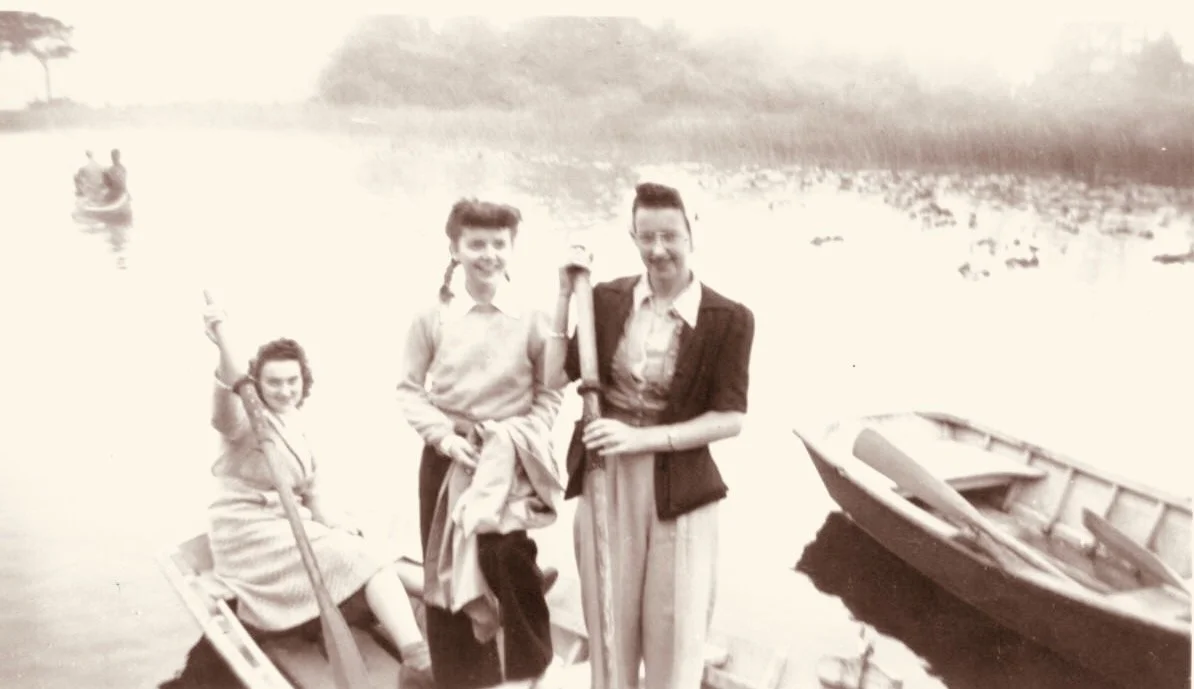 Three women standing near a boat on a river, with two women sitting in a boat and one woman standing. Two small boats are visible, and a person in a boat is in the background.
