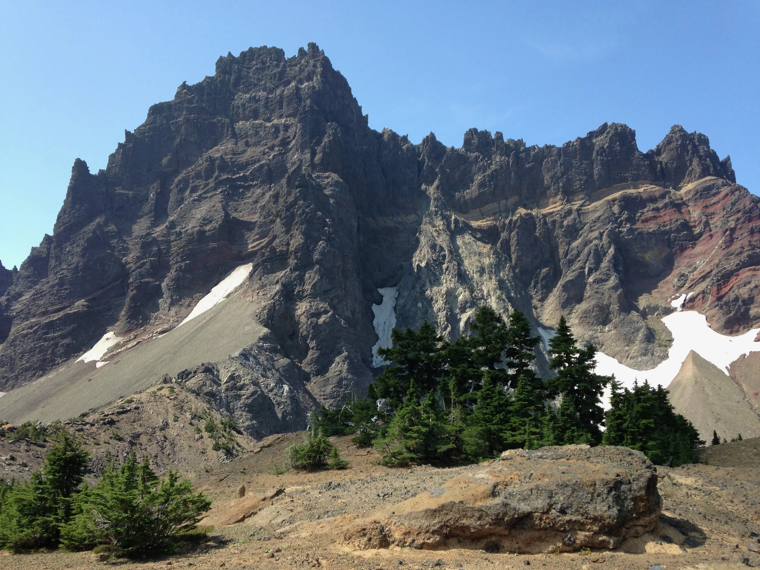 a rugged mountain with patches of snow and green trees in the foreground, under a clear blue sky