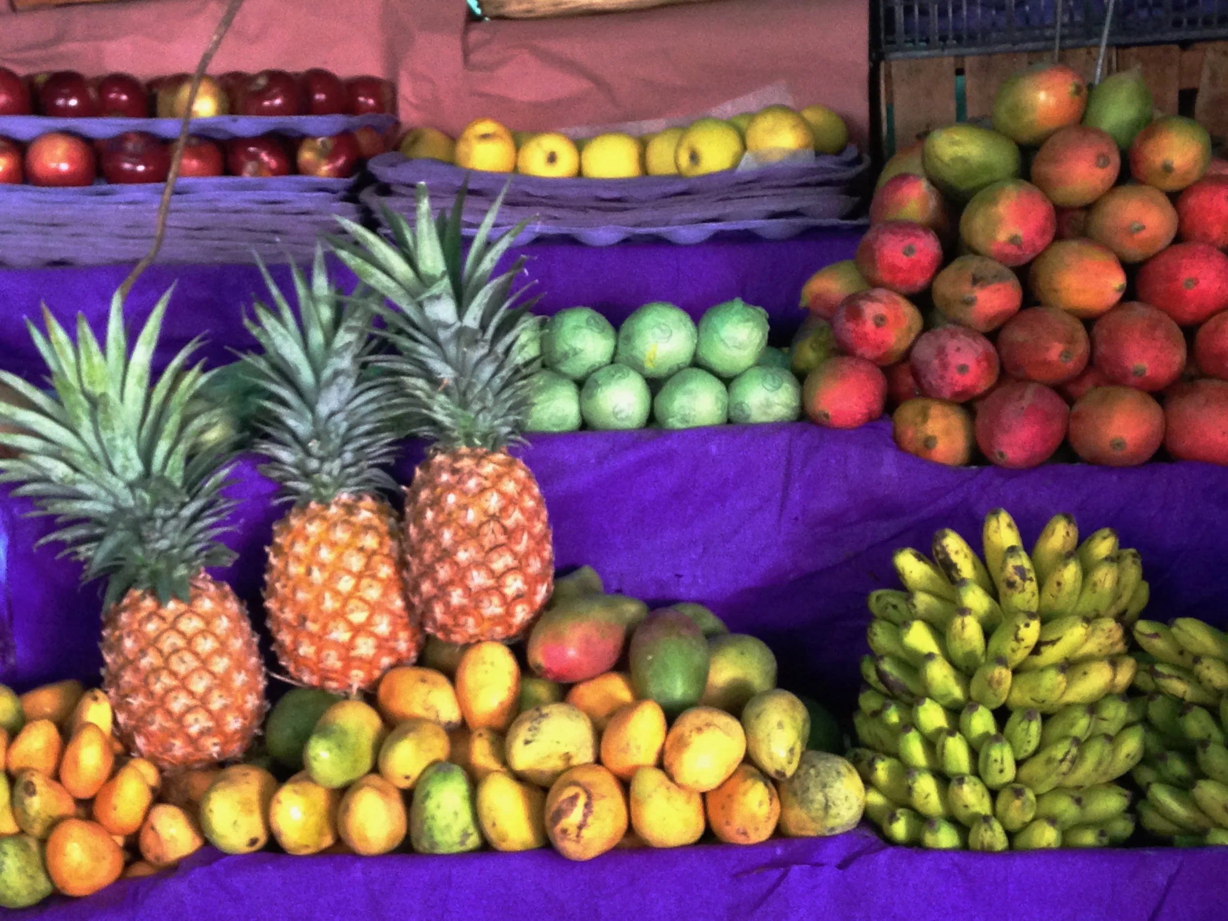 Fresh tropical fruits including pineapples, bananas, apples, mangoes, and pomegranates displayed at a market stall with purple cloth background.