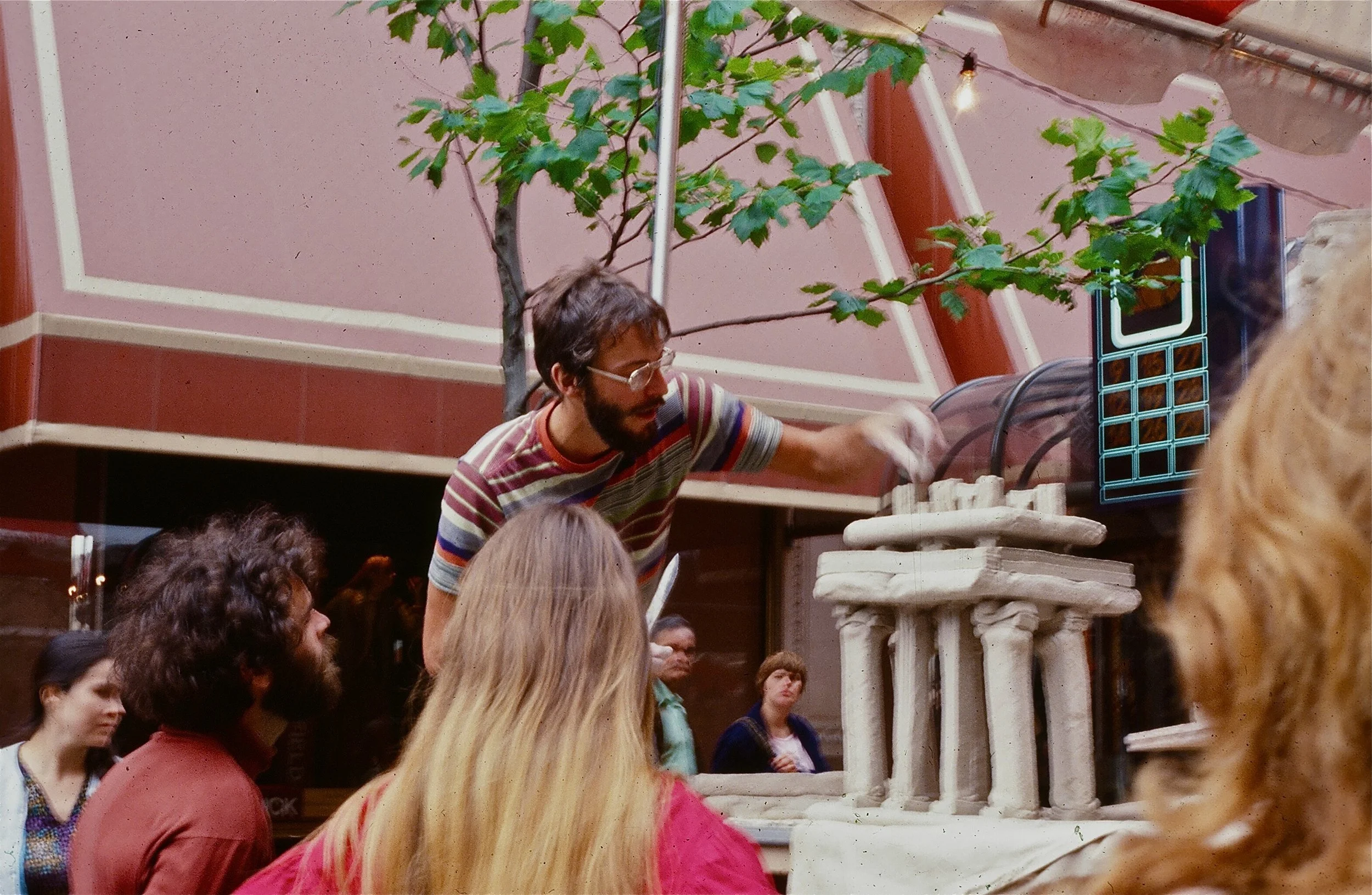 A man with glasses, a beard, and a striped shirt is demonstrating a scientific experiment to a group of people. The setup includes a structure resembling ancient columns, and there is a small tree and a pinkish building wall in the background.