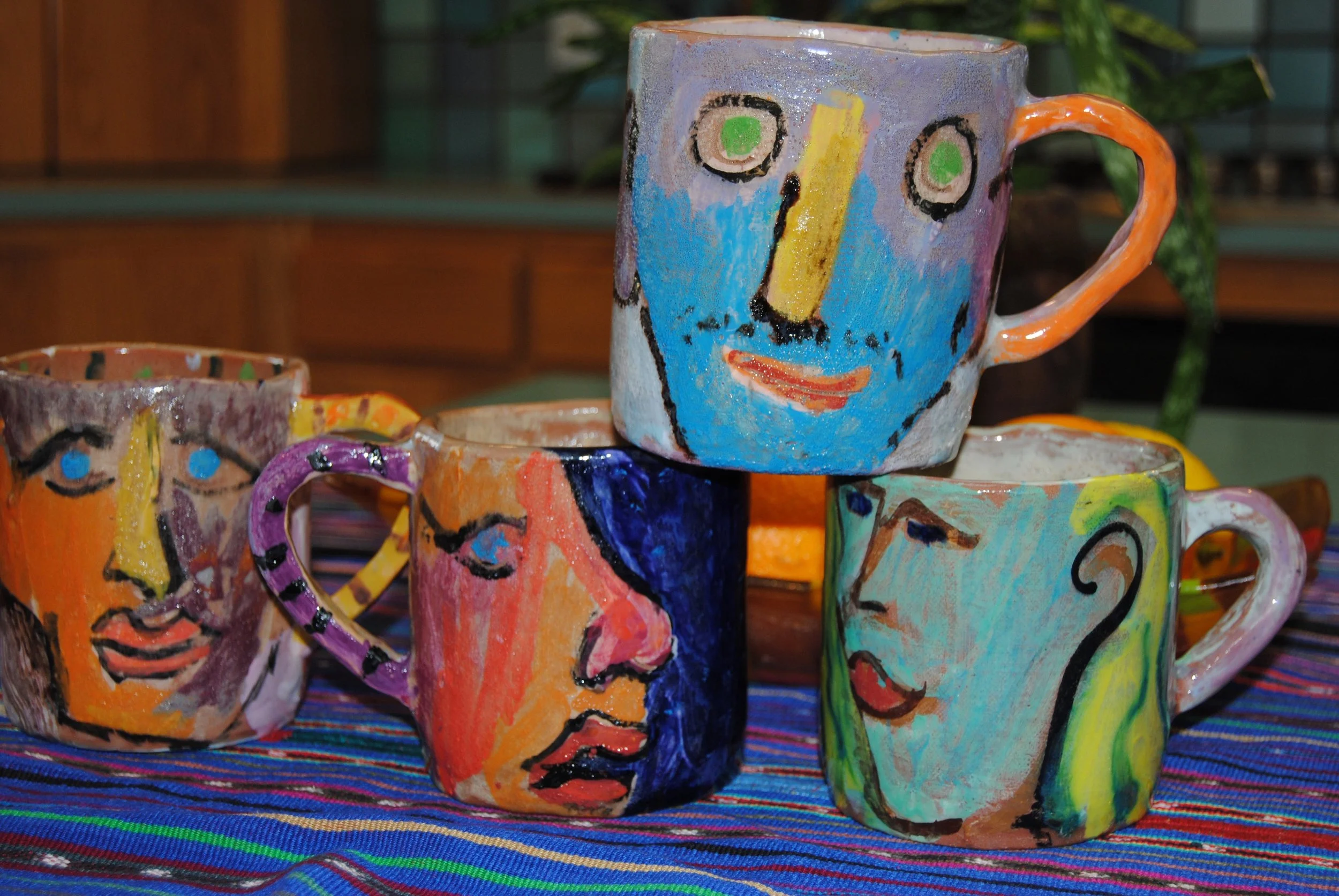 Colorful ceramic mugs decorated with abstract portrait paintings of faces, placed on a woven fabric with bright stripes.