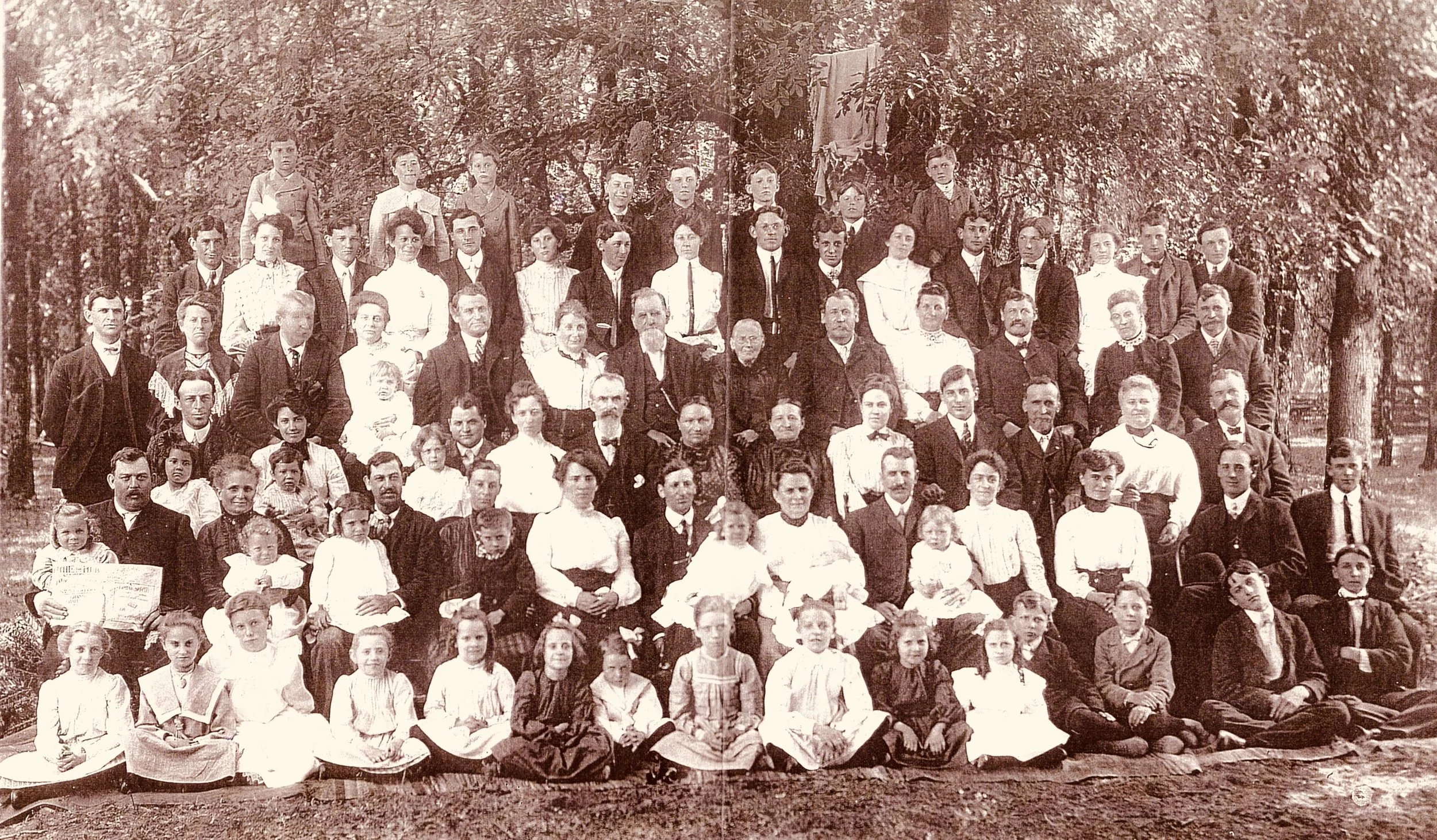 A large black-and-white photograph of a family group portrait outdoors during daytime, with numerous men, women, and children arranged in rows in front of trees.